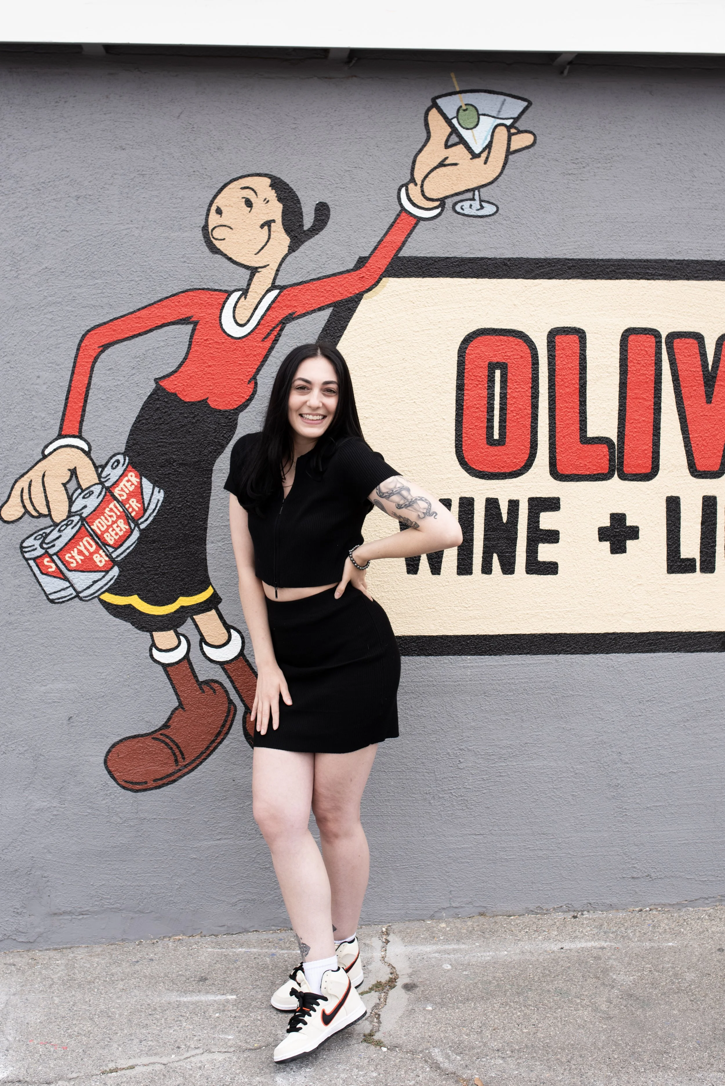 Young woman with black hair, wearing a black crop top, black skirt, and white sneakers with black laces, standing in front of a wall featuring a cartoon mural of Betty Boop holding a martini glass.