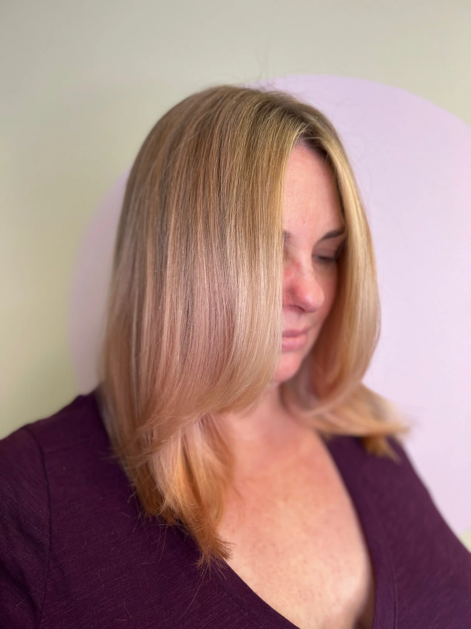 Close-up image of a woman with long, blonde hair wearing a purple top, with her eyes closed, against a light-colored background.