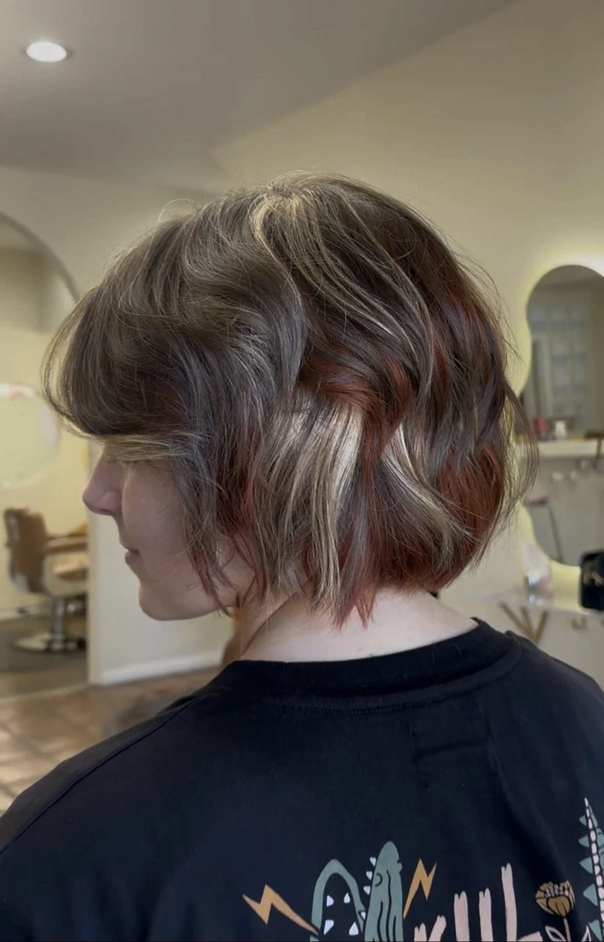 Side view of a woman with short, wavy hair that is blonde at the top and fades into reddish-brown and red at the bottom, in a salon or home setting.