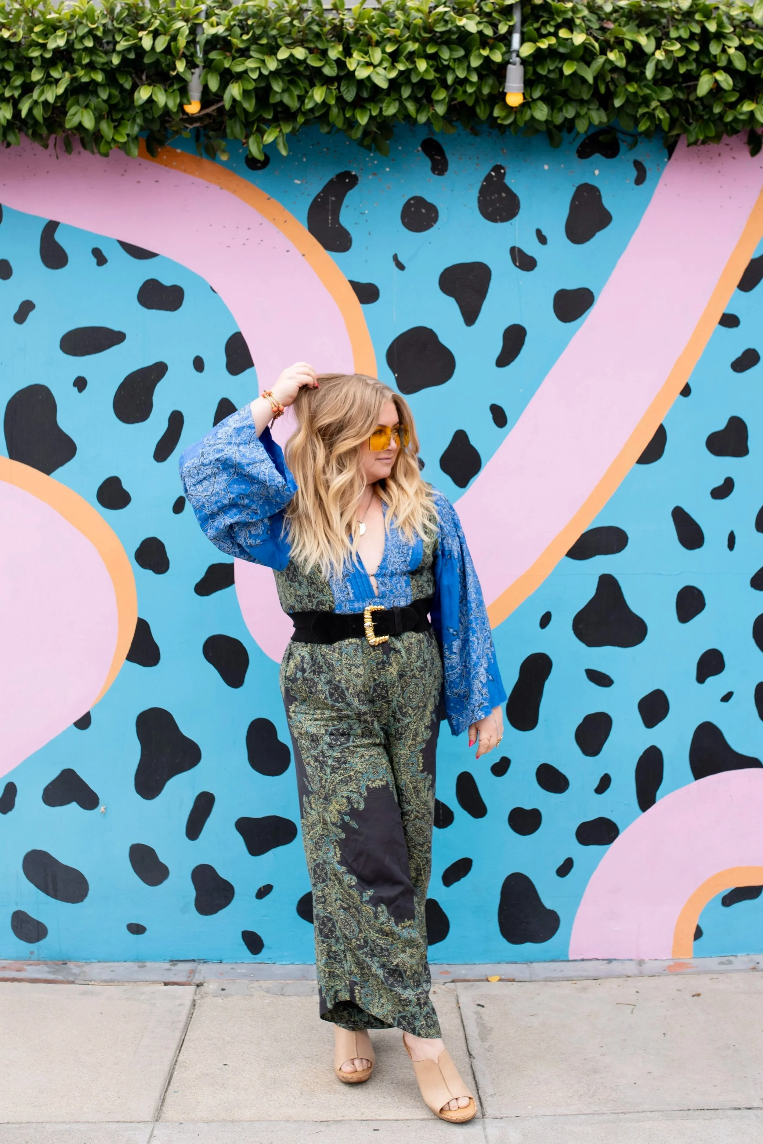 A woman with wavy blonde hair and yellow sunglasses stands in front of a colorful abstract mural with pink, black, blue, and orange shapes, wearing a patterned blue jacket and green patterned pants.