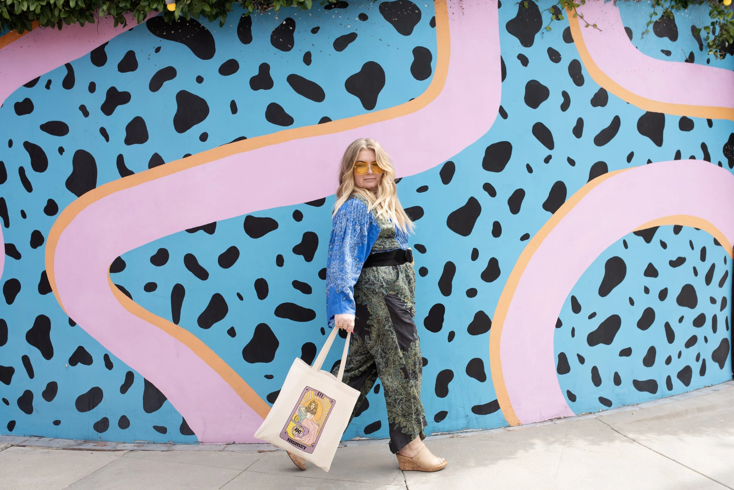 A woman standing in front of a colorful, abstract mural with pink, blue, black, and green shapes. She is wearing yellow sunglasses, a blue patterned jacket, patterned pants, and tan platform shoes, and is holding a tote bag with a tarot card design on it.