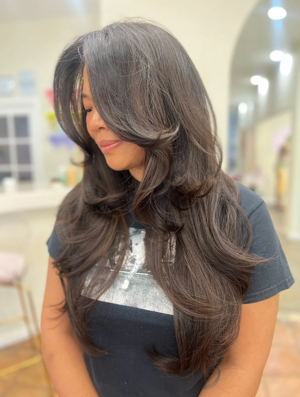 A woman with long, wavy brown hair with subtle highlights, smiling slightly, wearing a black t-shirt, in a softly lit indoor setting.