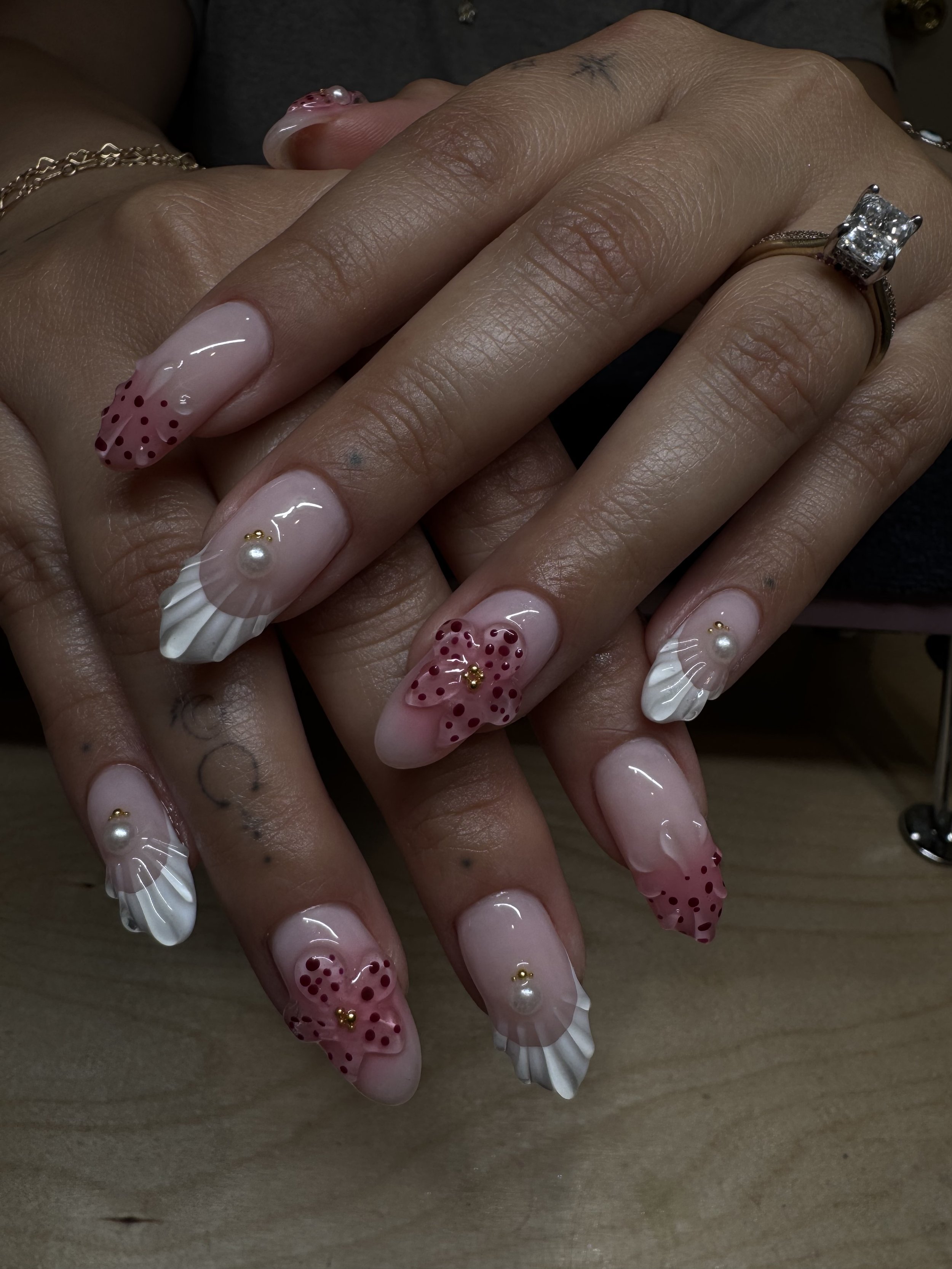 Close-up of hands with elaborately decorated acrylic nails featuring pink bows with red dots and pearl accents, jewelry including rings and bracelets.