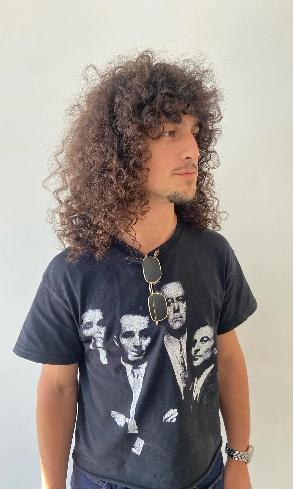 A man with curly hair wearing sunglasses hanging on his shirt and a black t-shirt with graphic images of four people, standing against a plain white background.