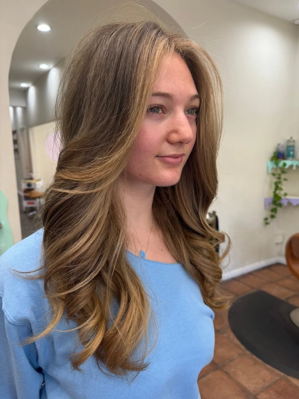 A young woman with long, wavy, light brown hair and subtle makeup, wearing a light blue top, standing indoors with a neutral expression.