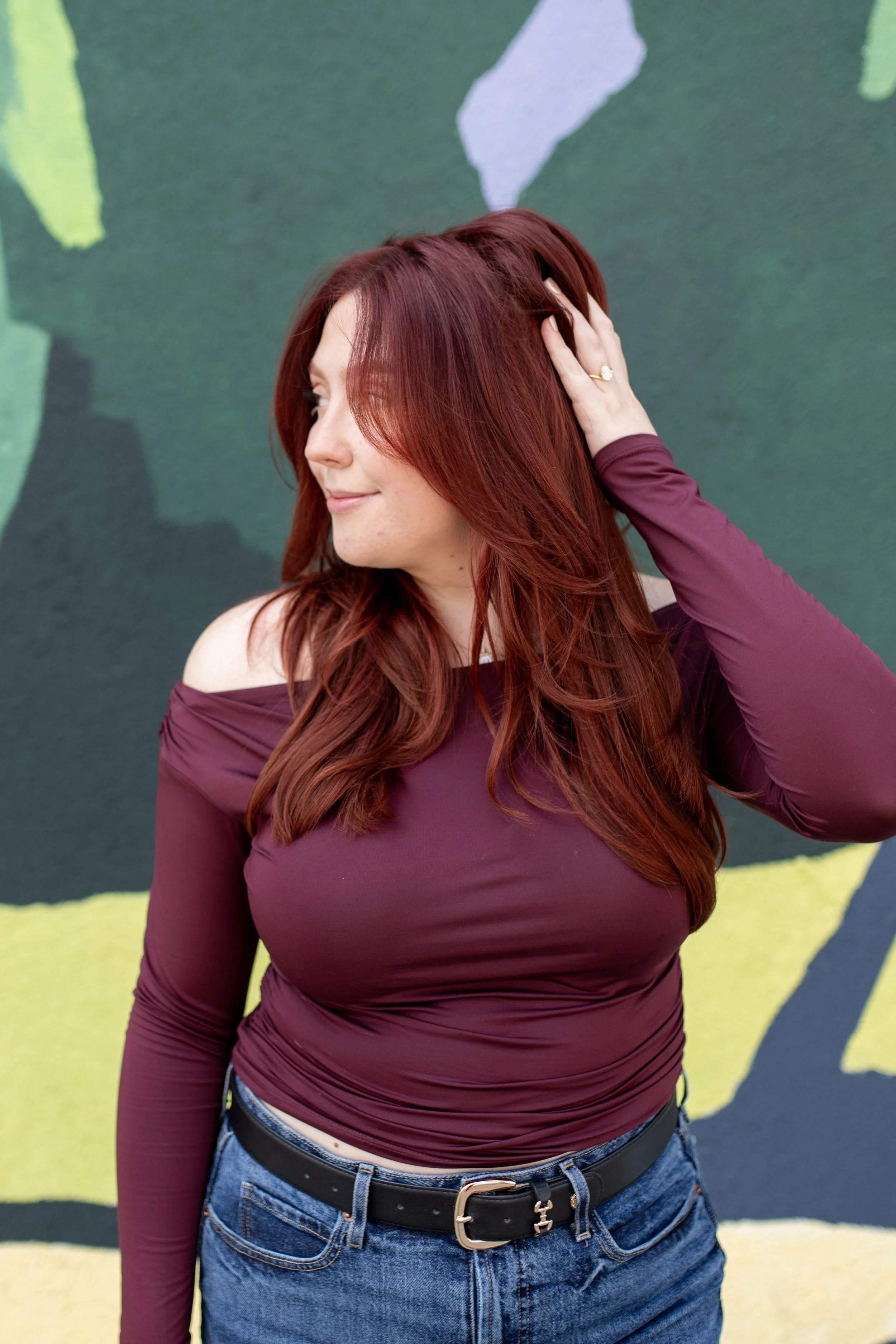A woman with long, wavy red hair wearing a purple long-sleeve top and blue jeans, standing against a colorful abstract background.