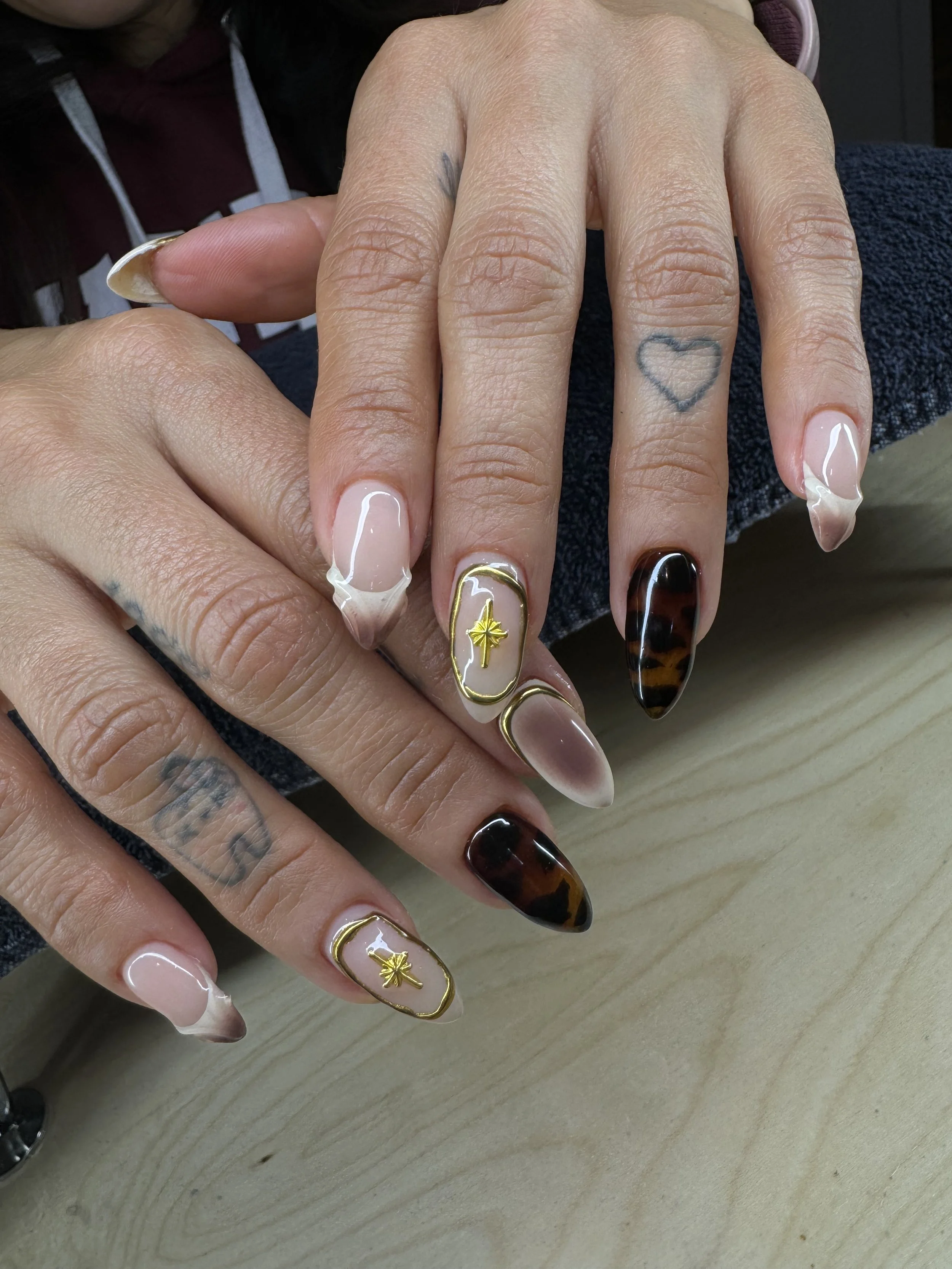 Close-up of a person's hands with decorated nails and small tattoos, including a heart on the middle finger, a star on the ring finger, and tattoos on the fingers. The nails are shaped long with decorative nail art.