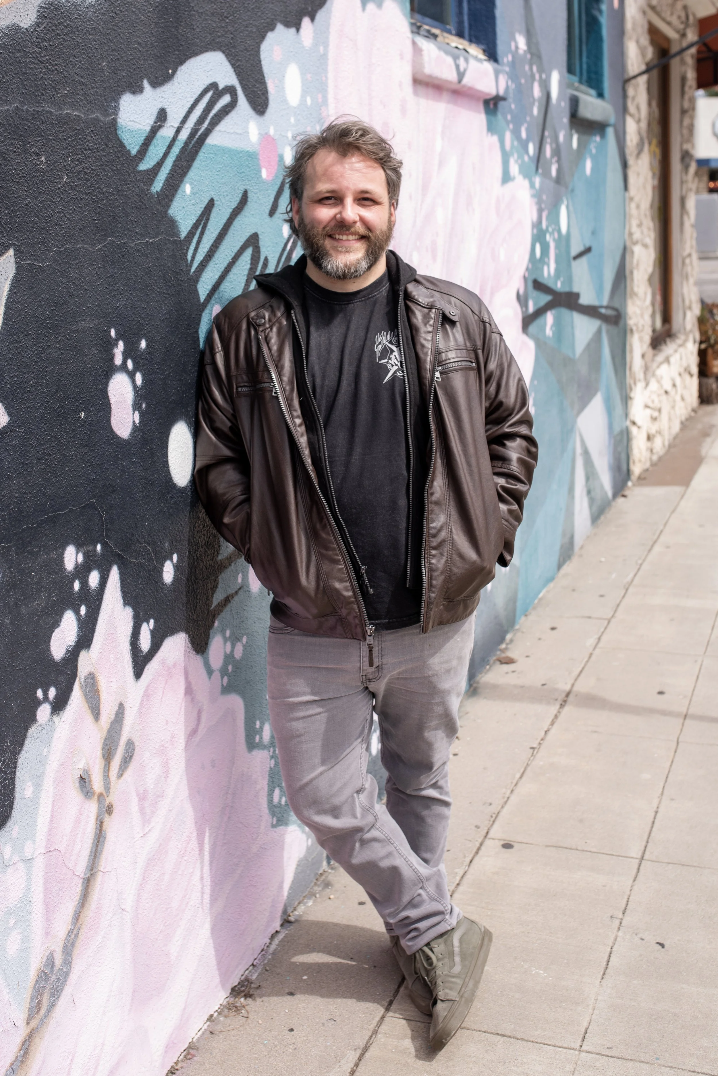 A man with a beard wearing a black jacket and gray pants, smiling and standing against a colorful mural on a city sidewalk.