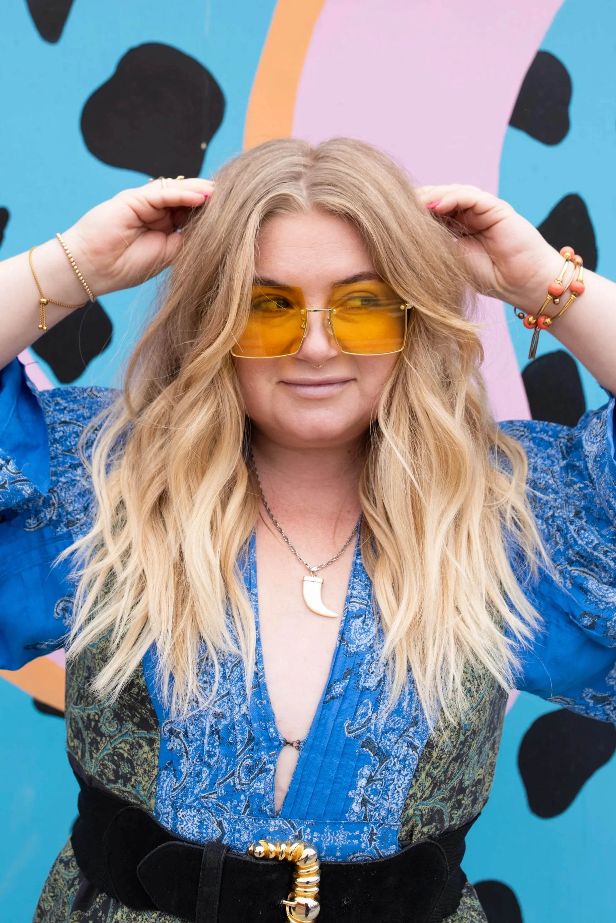 Nicoletta Gauci, long blonde hair wearing orange sunglasses and a blue patterned shirt, standing in front of a colorful, abstract background. She has bracelets and a necklace, and is adjusting her hair with both hands.