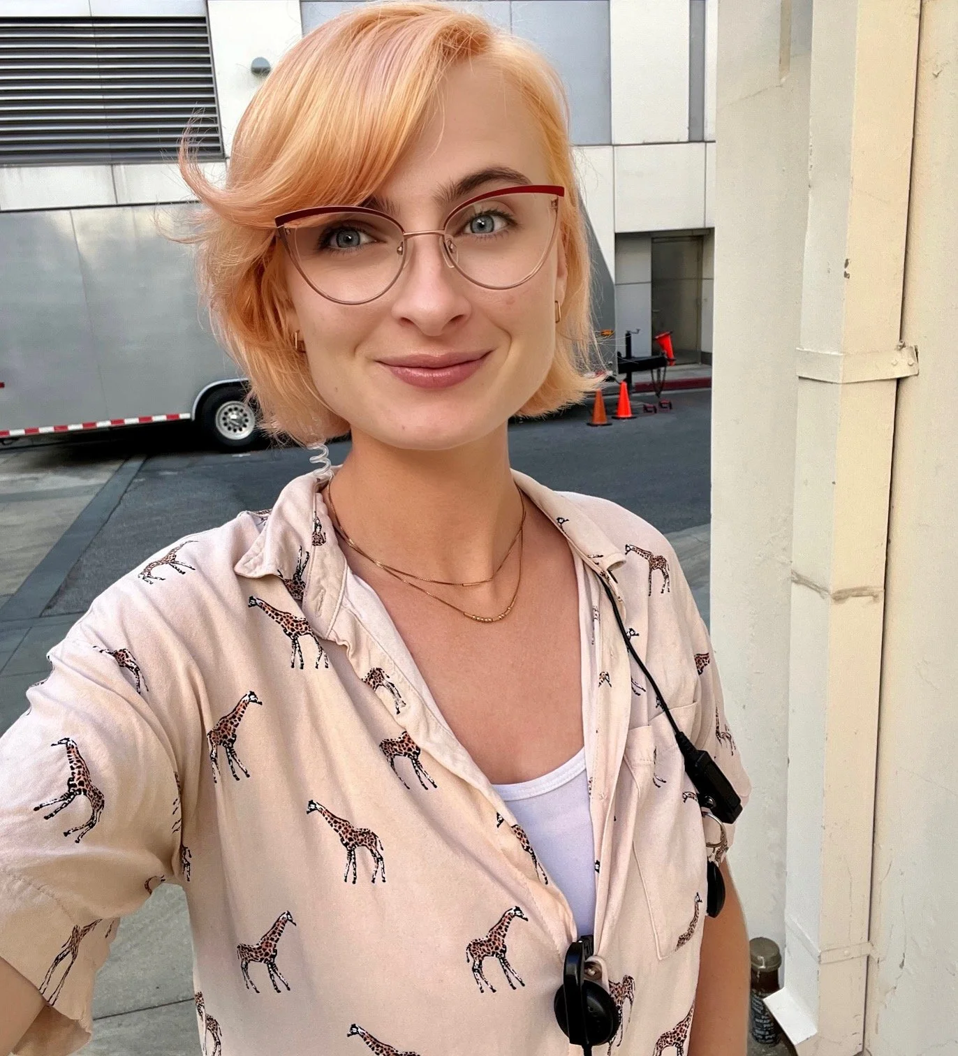 A young woman with short orange hair, glasses, and a light peach shirt with giraffes, standing outside near a building with orange cones and a trailer in the background.