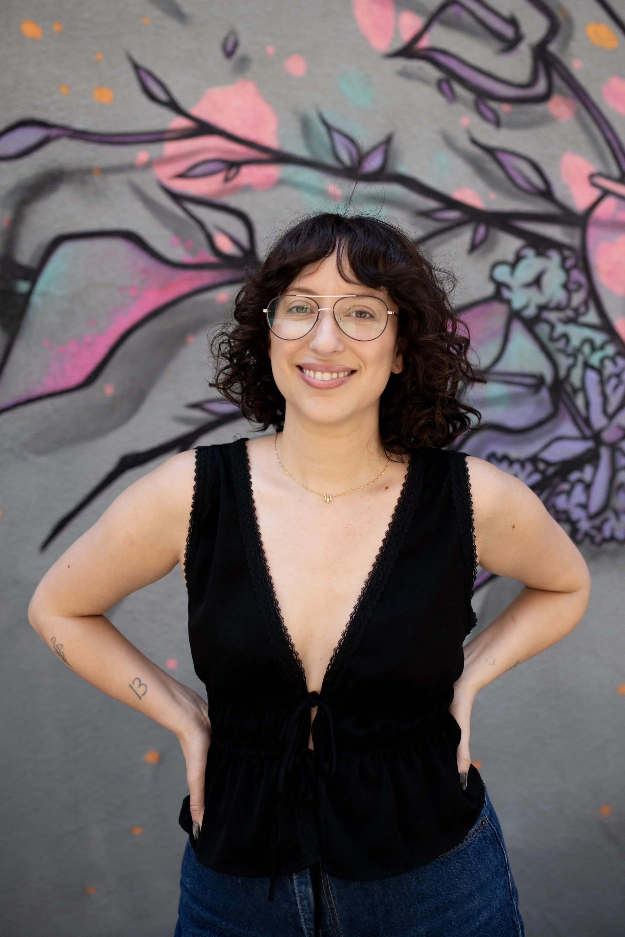 A smiling woman with curly dark hair and glasses standing in front of a colorful mural on a gray wall. She is wearing a black sleeveless top and blue jeans.