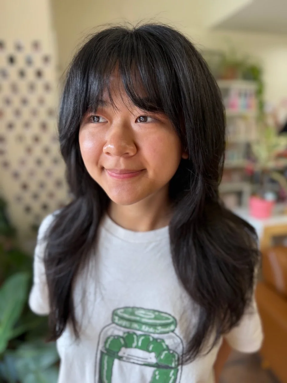 A young woman with long, dark hair and bangs, smiling softly in a cozy indoor setting with a blurred background of books and plants.