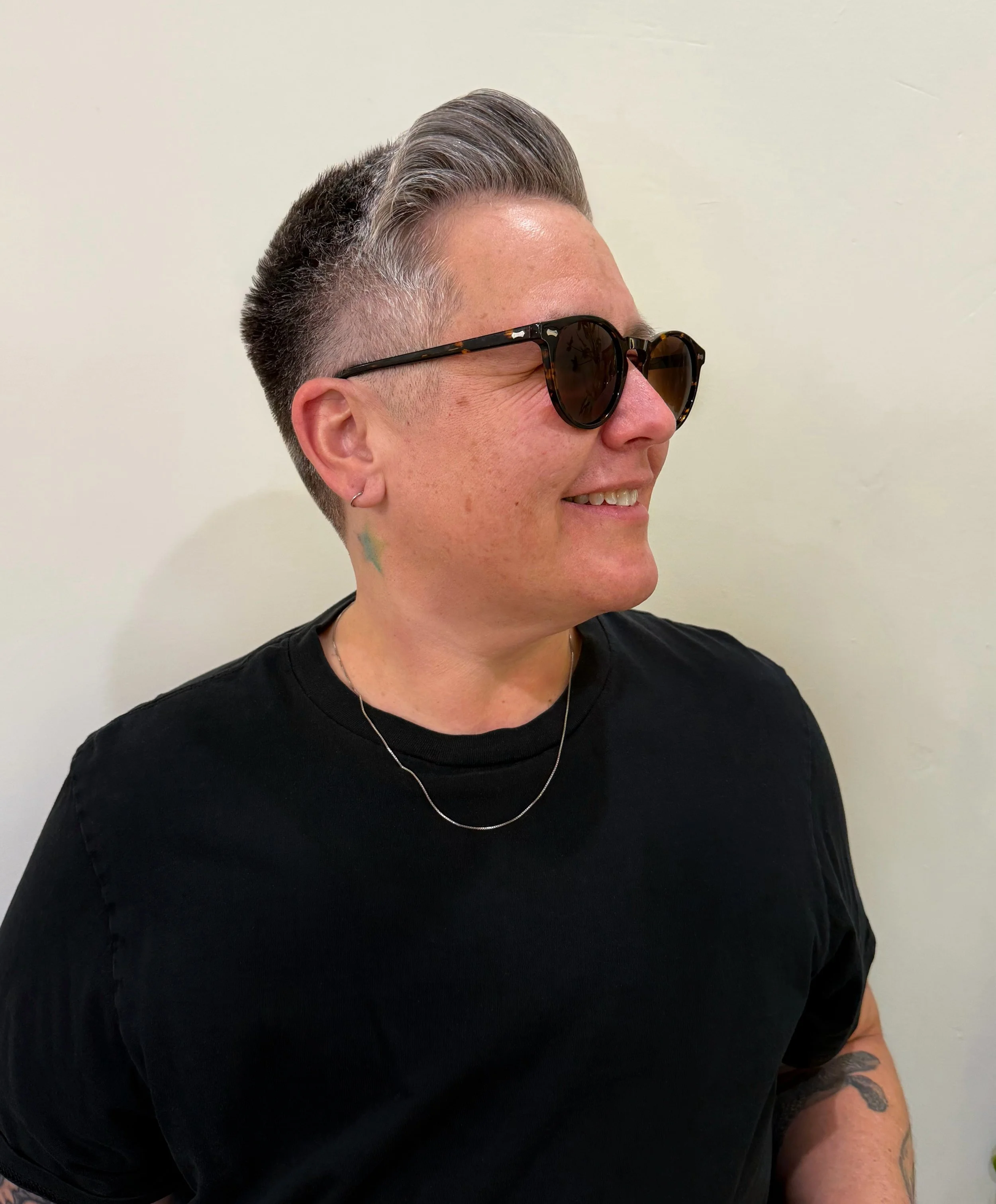 A person with short gray hair, wearing tortoise-shell sunglasses, a black shirt, and a silver necklace, smiling slightly, against a light-colored wall.
