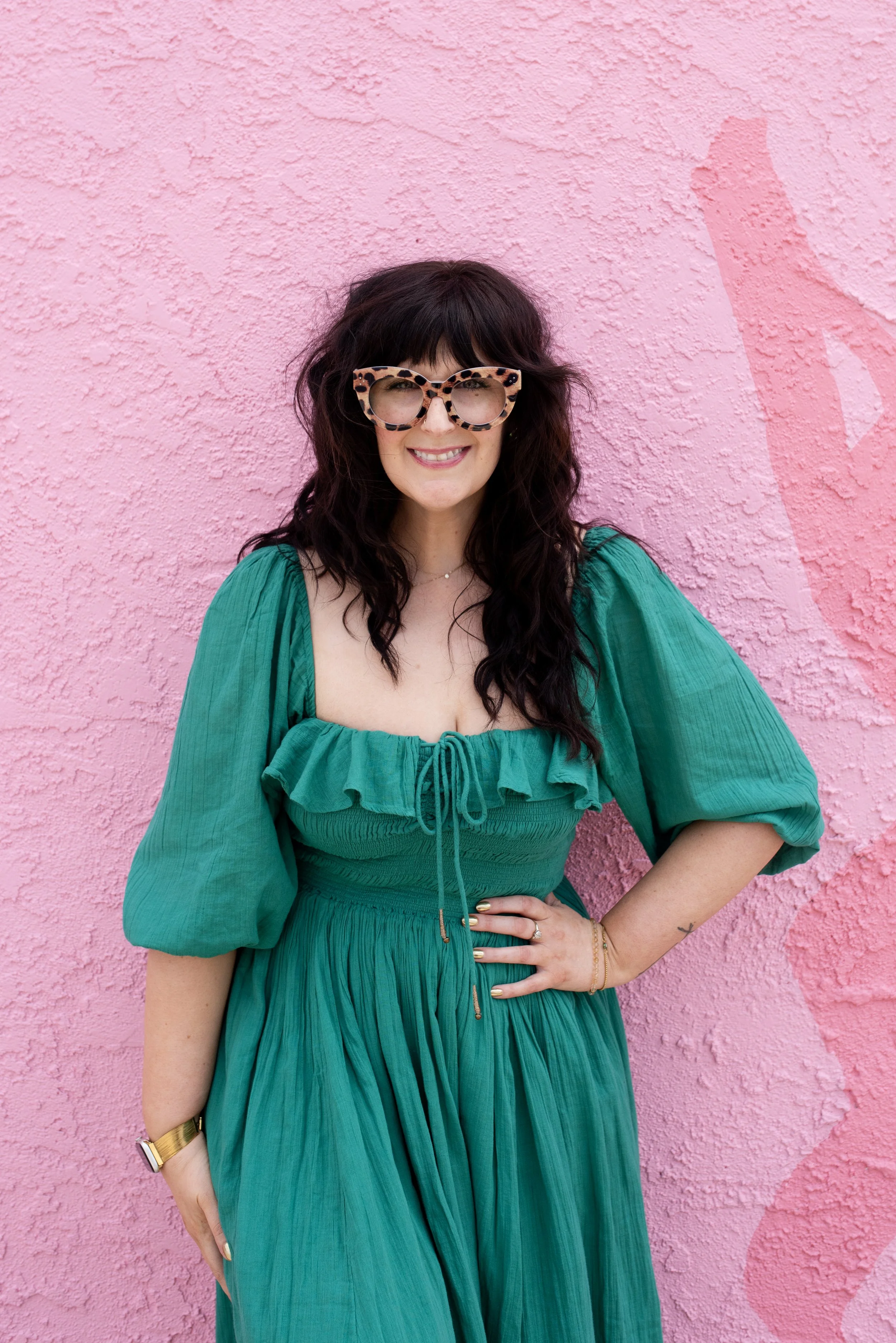 A woman with long dark hair and tortoiseshell glasses smiling and wearing a green dress, standing in front of a pink textured wall.