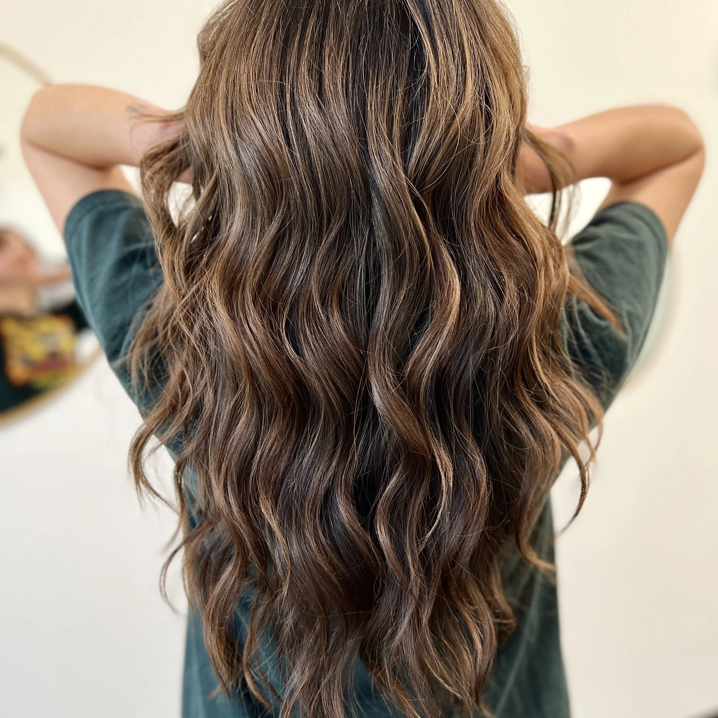 Back view of a woman with long, wavy, brown hair, wearing a dark-colored top, standing against a plain, light-colored wall.