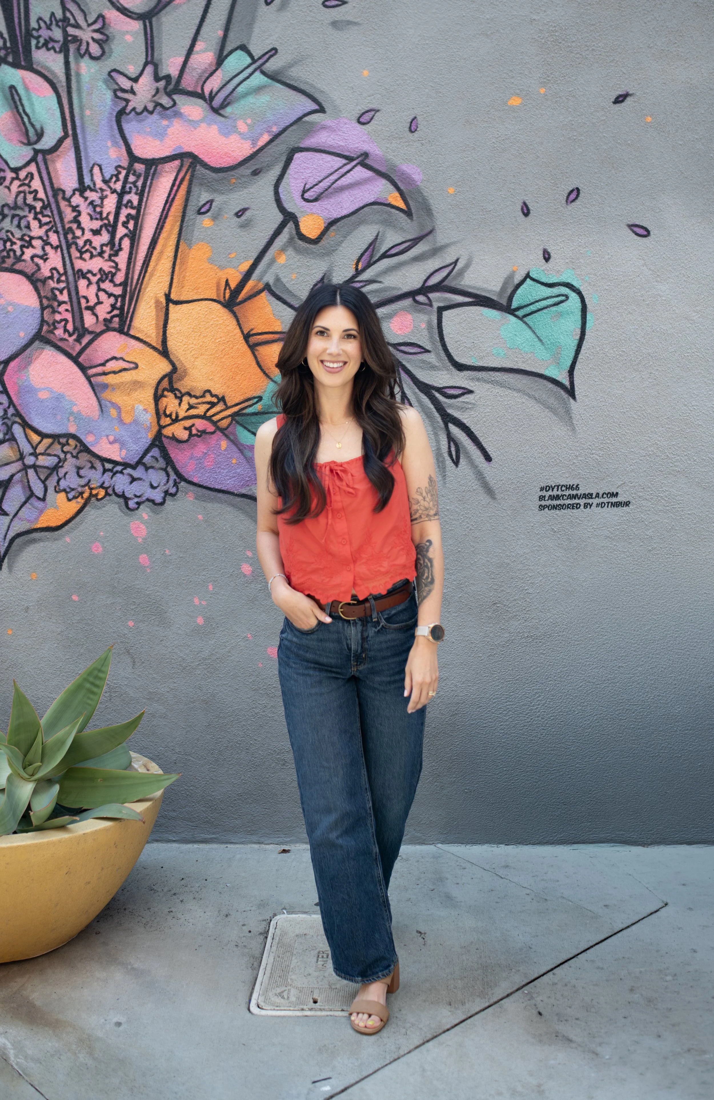 A woman with long dark hair in a red sleeveless top and blue jeans stands next to a large yellow pot with a green succulent plant. She is smiling and standing on a sidewalk in front of a colorful graffiti wall.