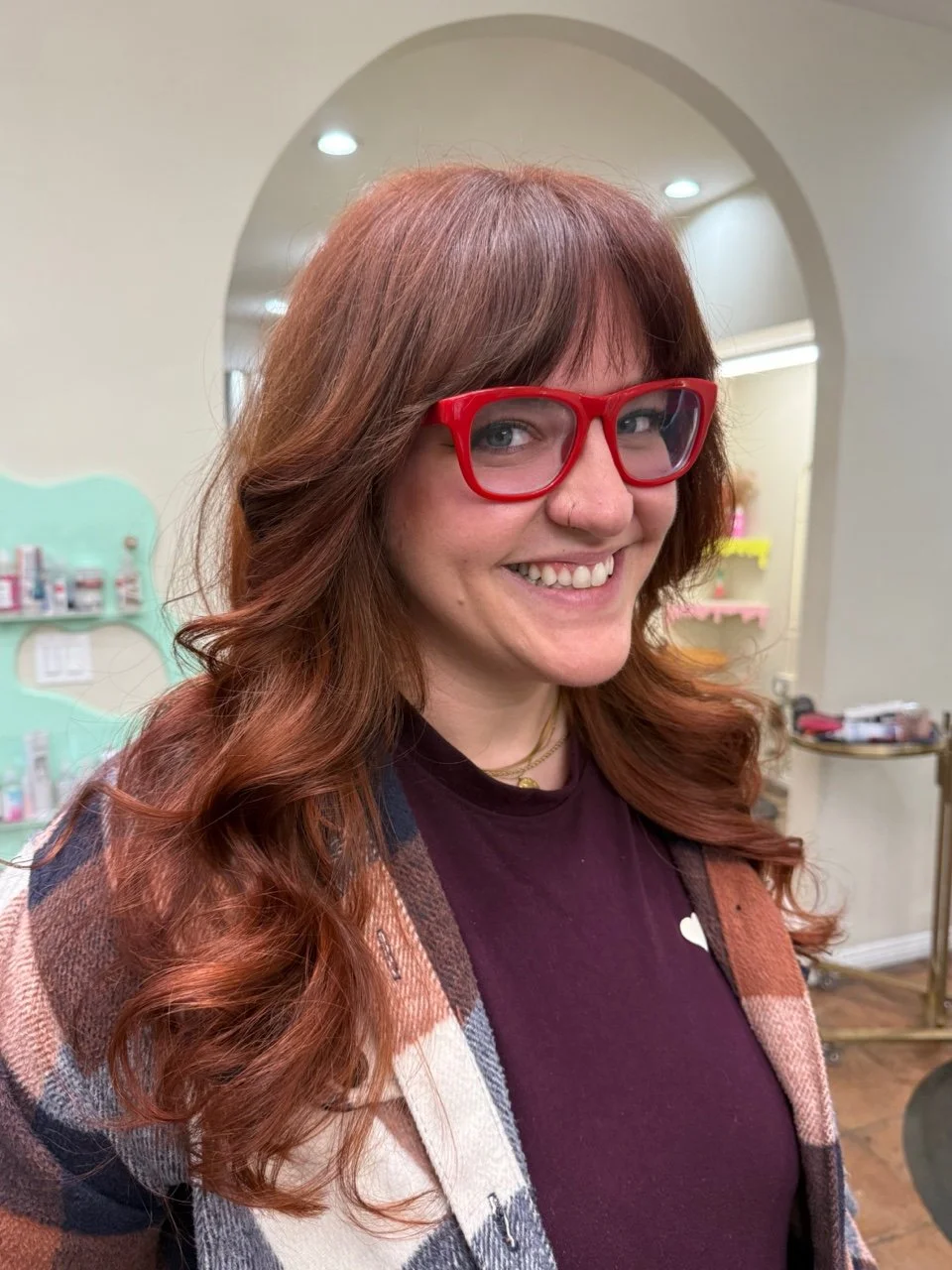 A smiling woman with red hair wearing red glasses, a maroon shirt, and a plaid jacket in a salon.