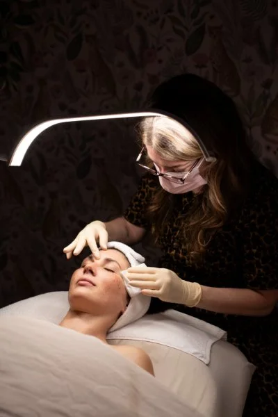 A woman with glasses and gloves performs a facial treatment on a woman lying in a bed with a towel wrapped around her head under a bright light.