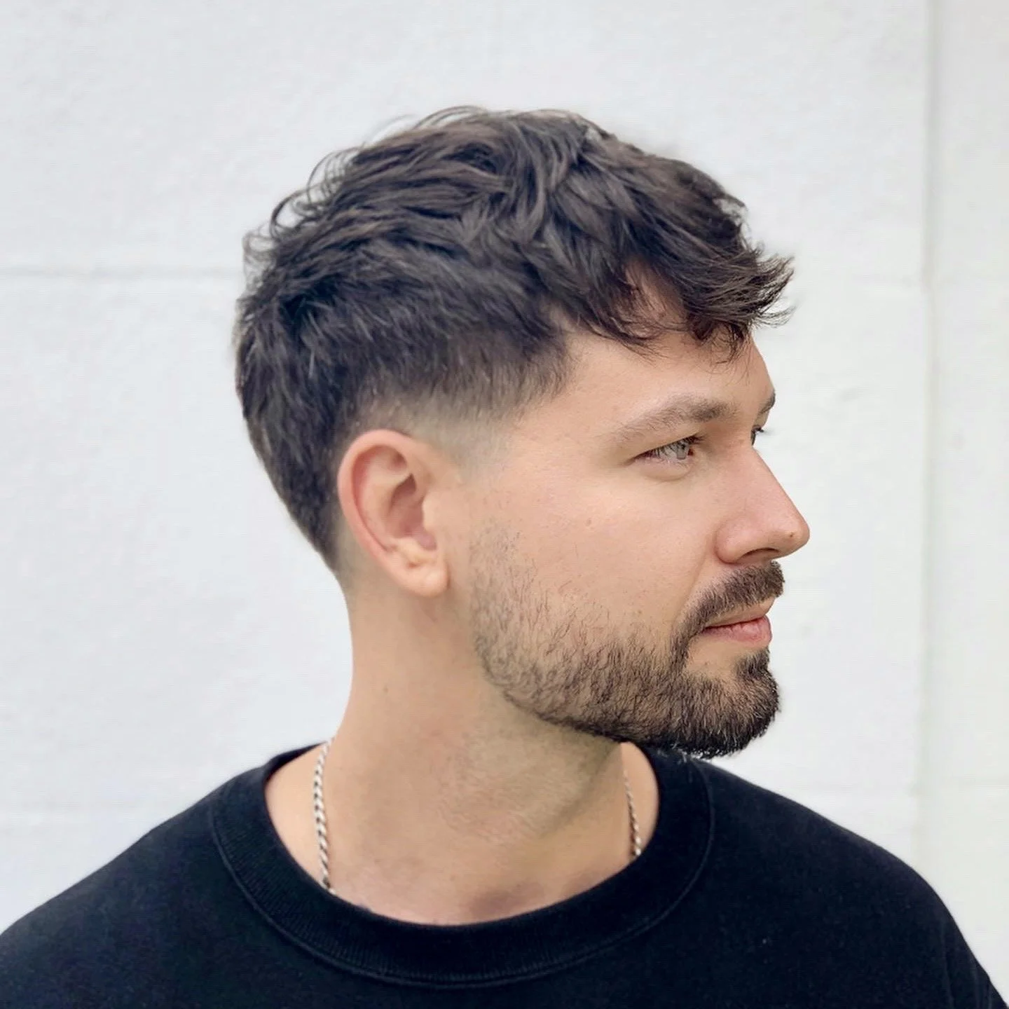 Side profile of a young man with styled hair, beard, wearing a black T-shirt and a silver chain, standing against a plain white wall.