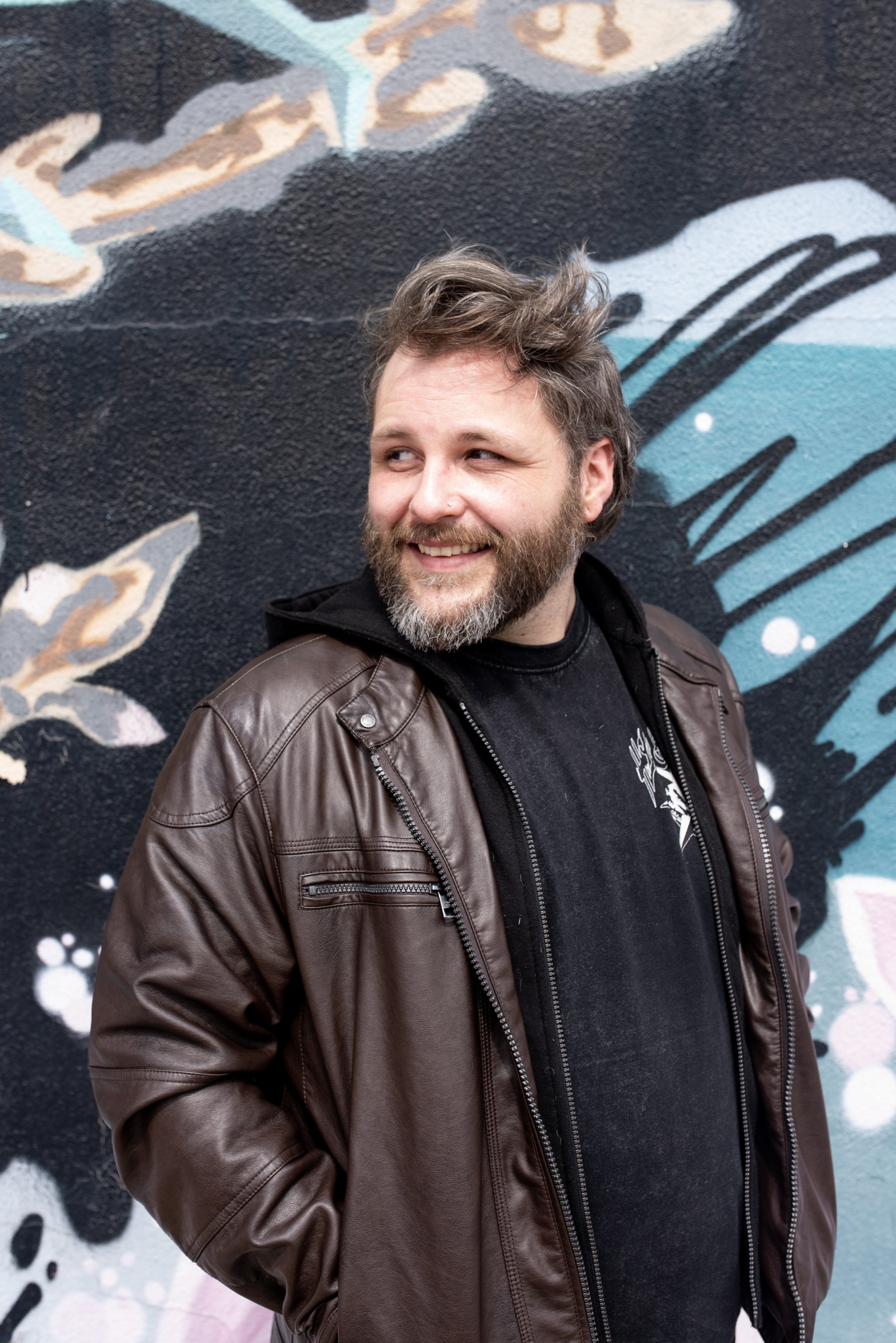 Danny Jouglet, a man with brown hair, a beard, and a nose piercing smiling while standing in front of a colorful mural wall.