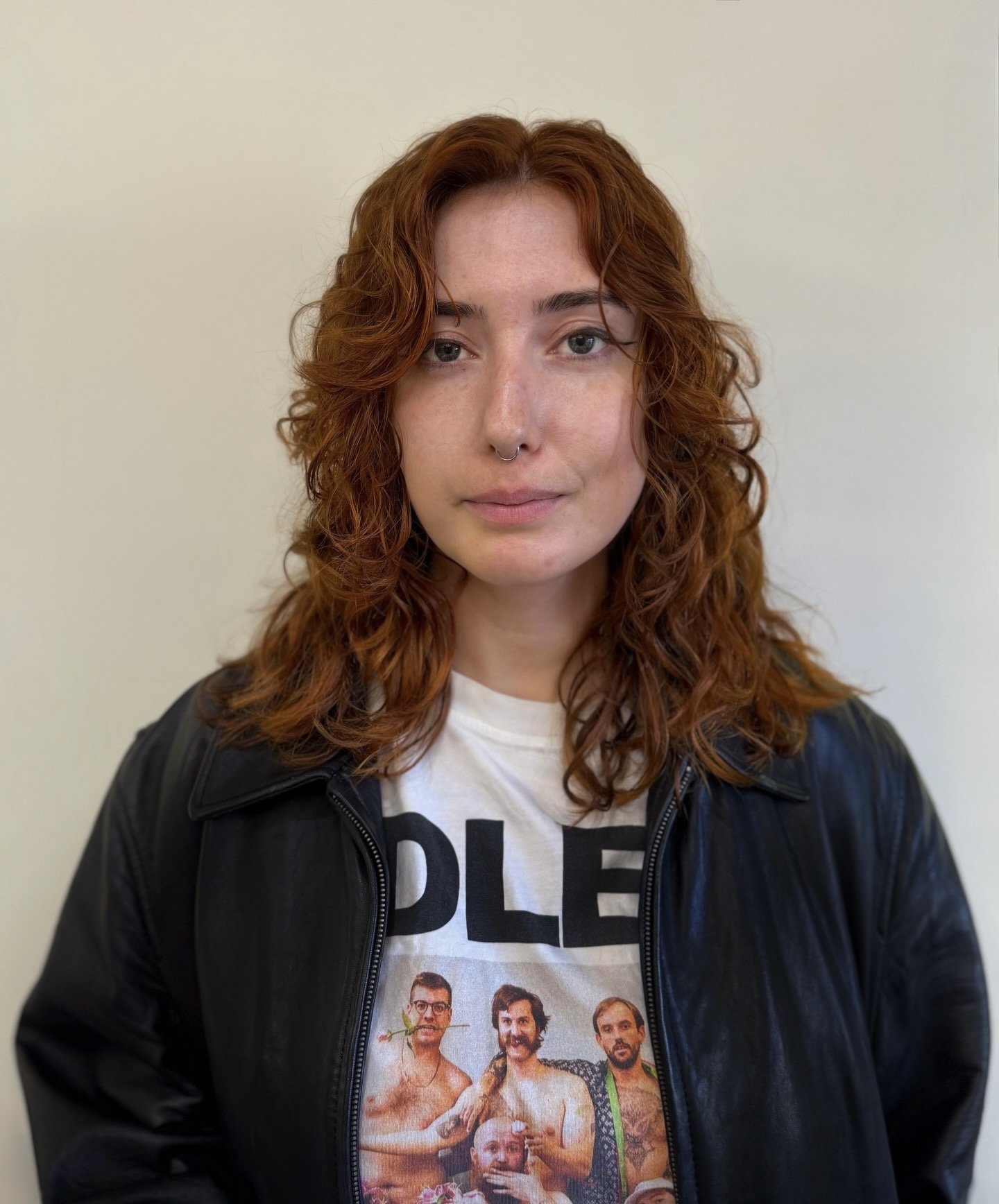 Young woman with shoulder-length curly red hair, wearing a black leather jacket and a graphic t-shirt, standing against a plain light-colored background.