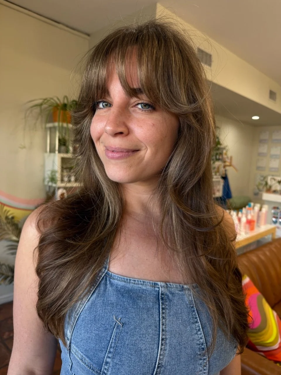 Close-up of a woman with long, wavy brown hair and bangs, smiling slightly, wearing a sleeveless denim top, in an indoor setting with plants and shelves in the background.