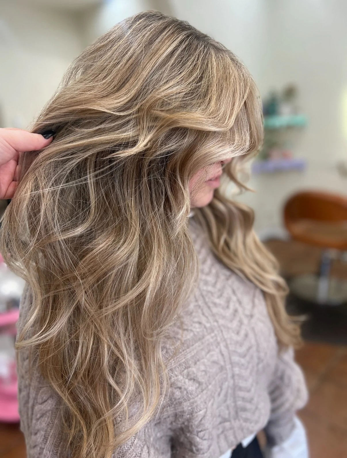 A woman with long, wavy blonde hair with highlights, wearing a beige textured top, is sitting in a salon. Someone is holding her hair to the side, showcasing its color and style.