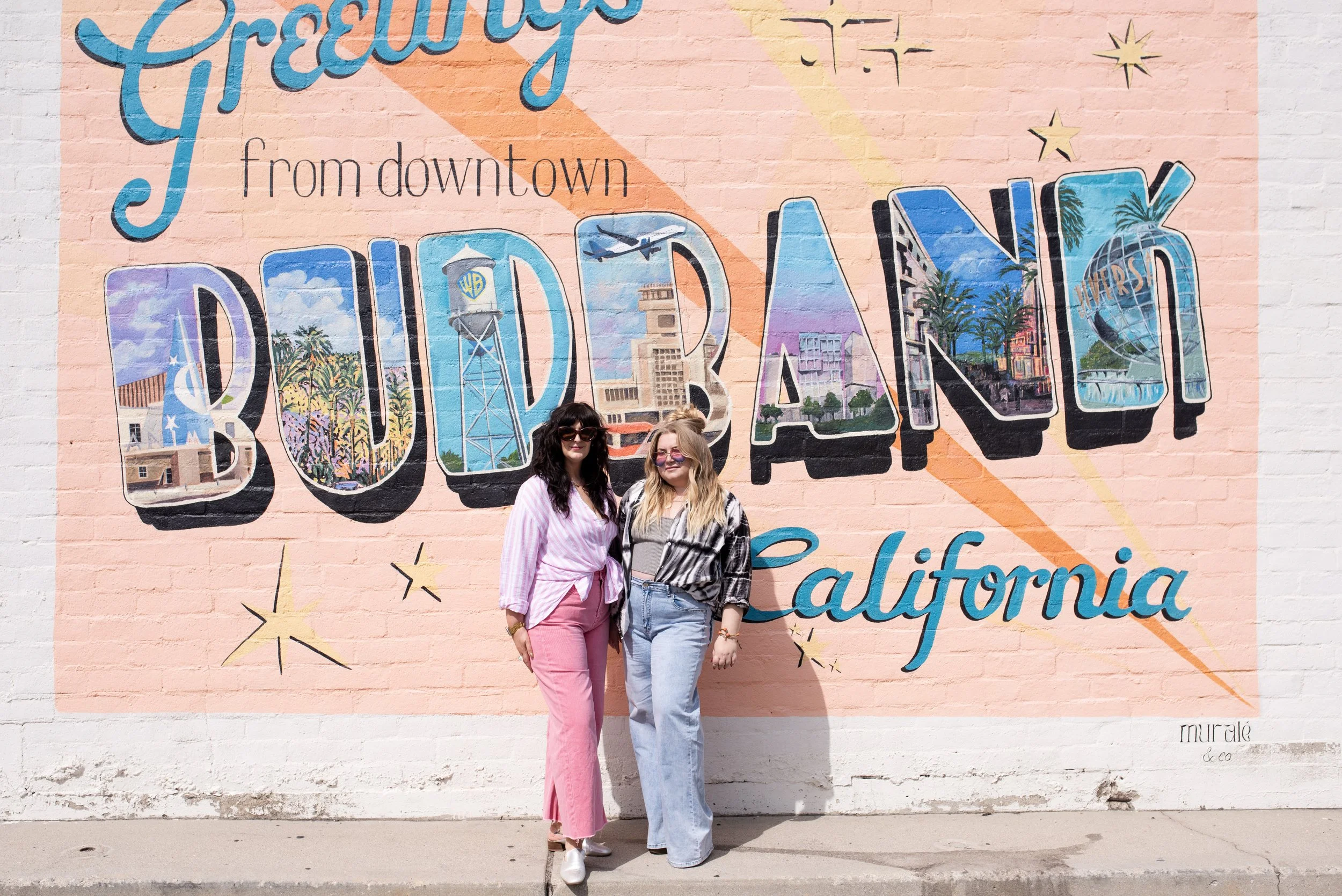 Two women stand in front of a colorful mural on a pink brick wall that reads 'Greetings from downtown California.' They are smiling, with the woman on the left wearing pink pants and sunglasses, and the woman on the right wearing light blue jeans and a black and white patterned shirt.