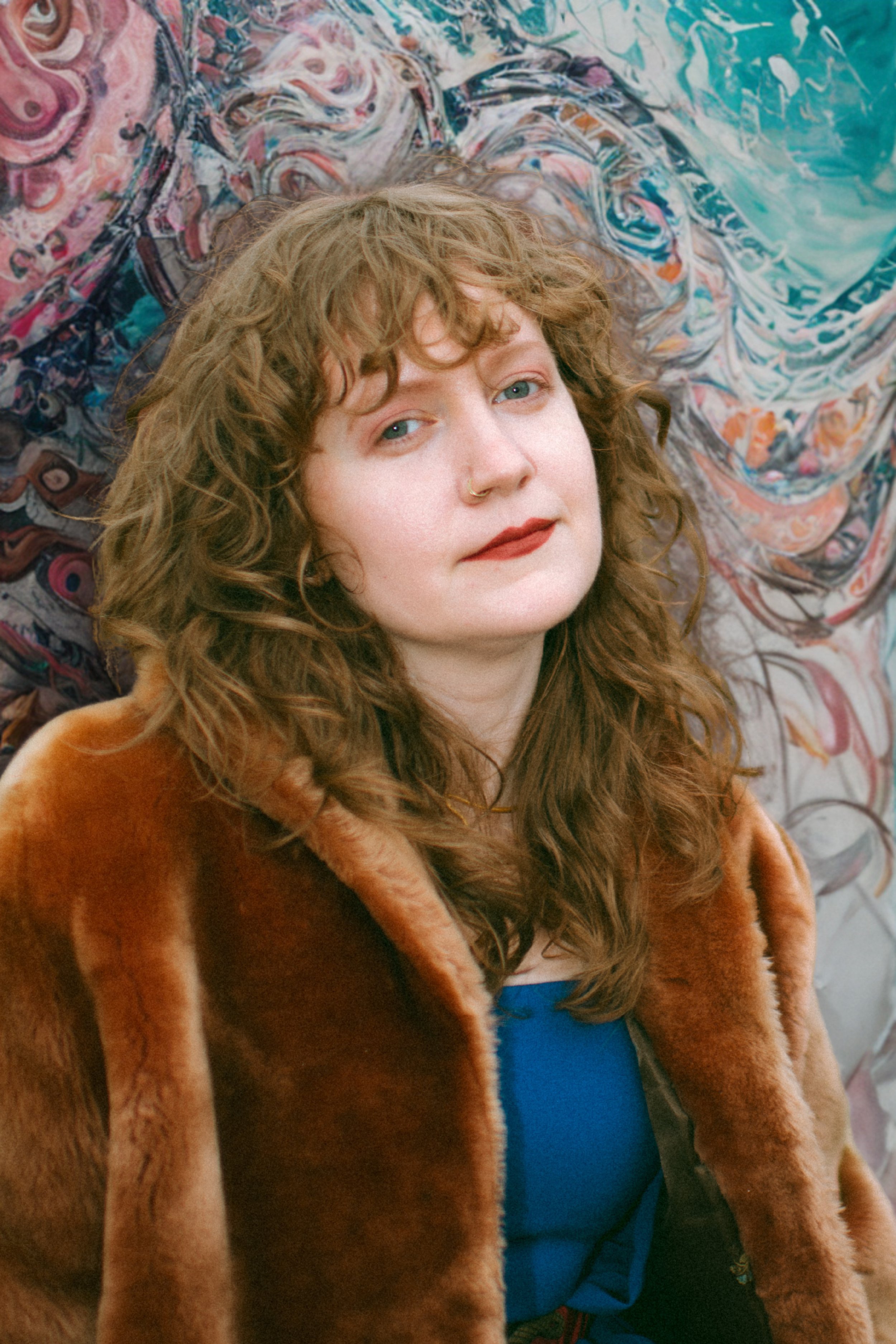 A woman with curly brown hair, wearing a fur coat and a blue shirt, lying down with her head turned slightly, looking directly at the camera. She has a nose ring and a calm expression. Behind her is a colorful, abstract patterned fabric.