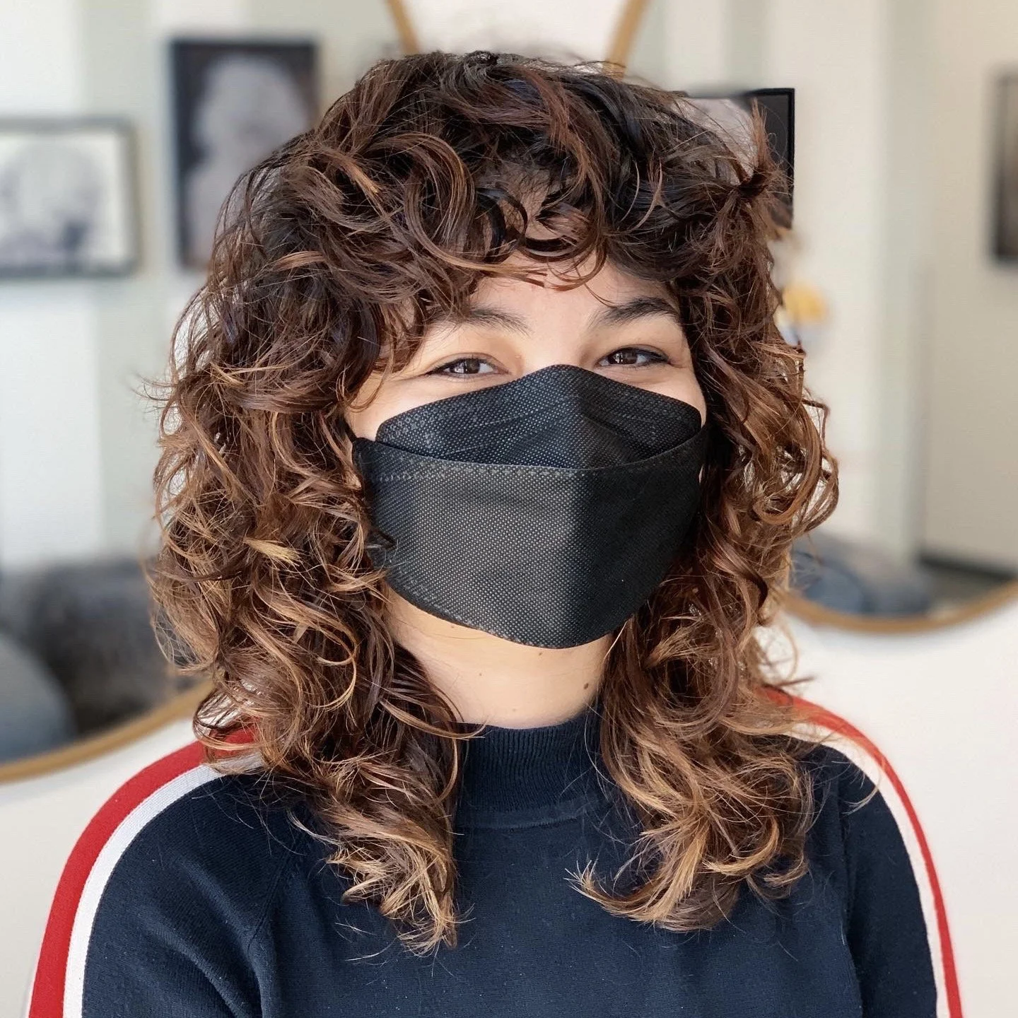 A woman with curly brown hair wearing a black face mask and a dark blue shirt with red and white stripes on the sleeves, standing indoors.