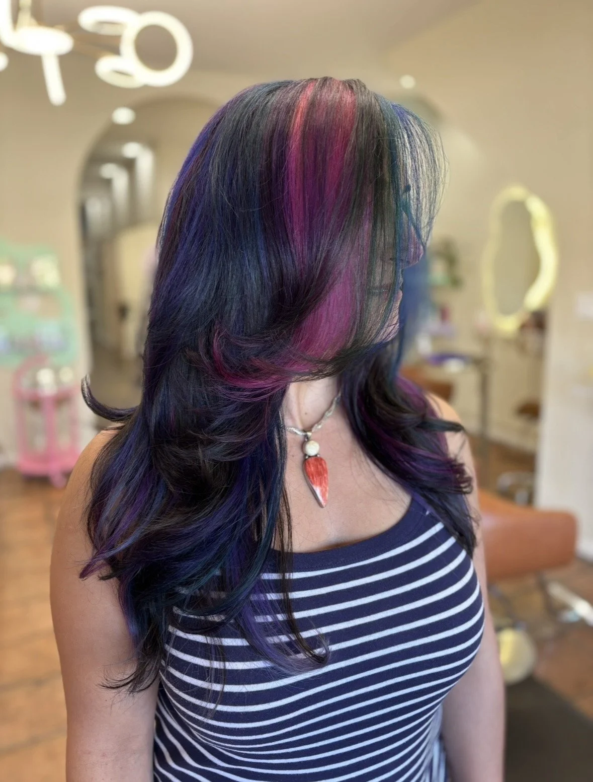A woman with long, wavy hair dyed in multiple colors, including purple, blue, and pink, wearing a striped tank top and a necklace with a red and white pendant, standing inside a salon.