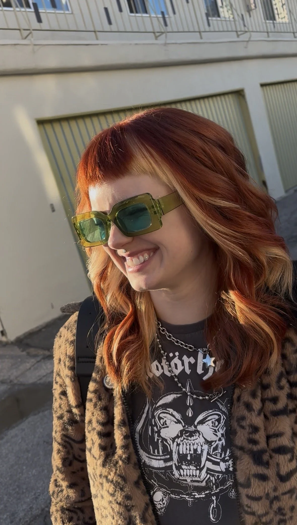 A young woman with reddish-orange hair, wearing green tinted sunglasses, a black graphic t-shirt, a leopard print coat, and layered jewelry, smiling outdoors during sunset.