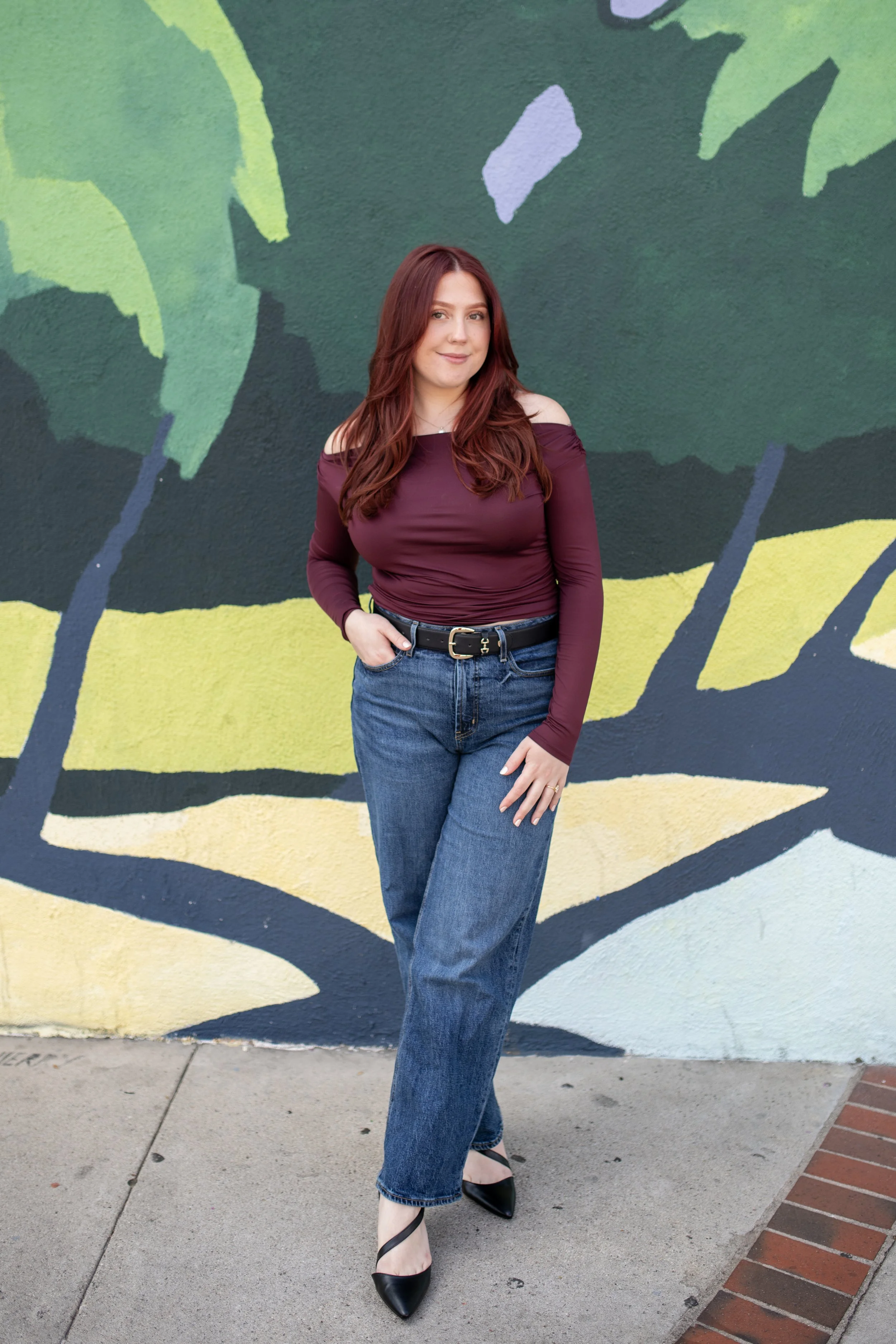 A woman with long, wavy, auburn hair stands in front of a colorful outdoor mural featuring green leaves and black branches. She wears a maroon off-shoulder top, blue jeans, and black pointed heels. She has a subtle smile with her right hand in her pocket.