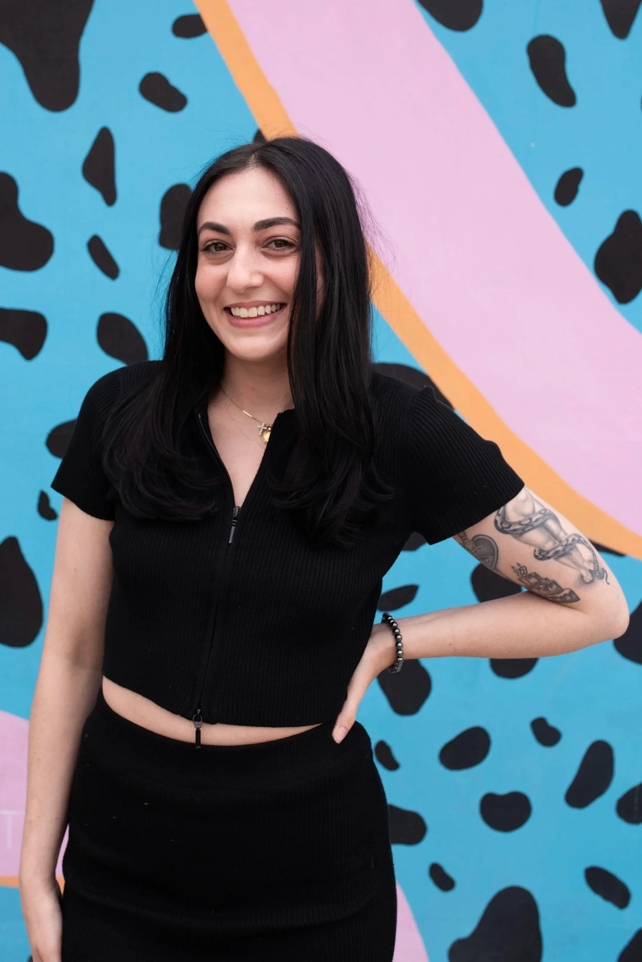 A young woman with long black hair, wearing a black zip-up top and jewelry, smiling while posing with a colorful, abstract graffiti background.