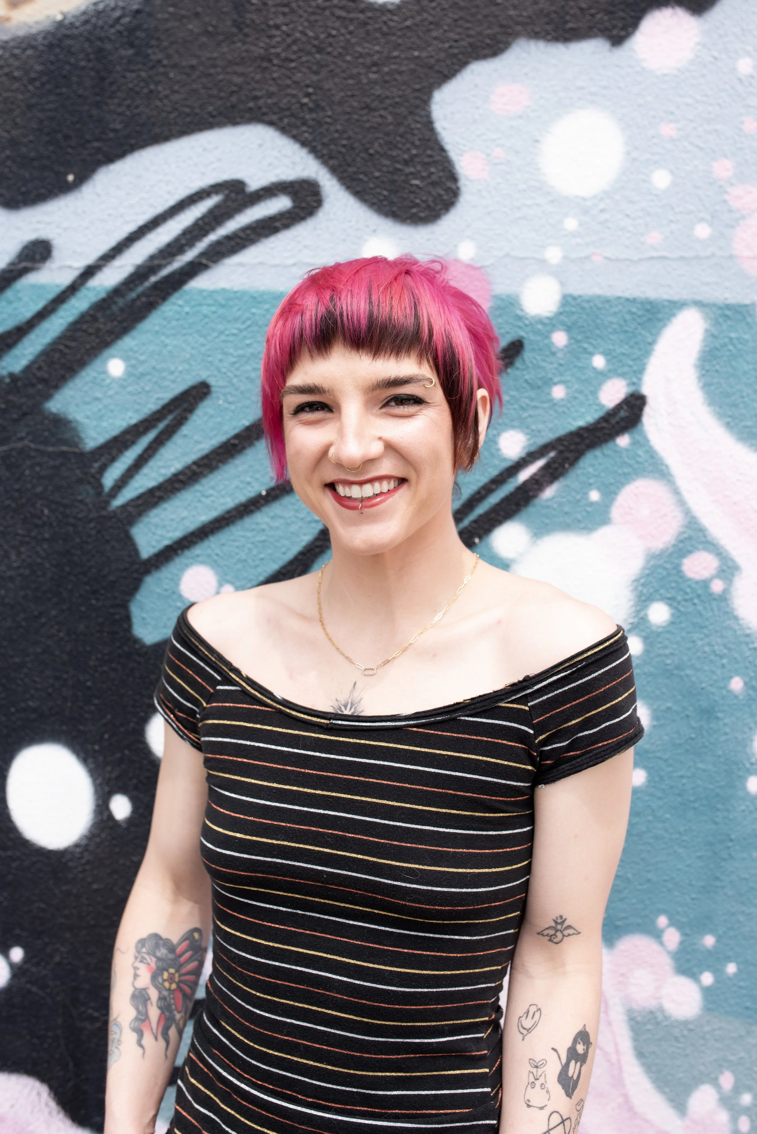 A woman with pink and black hair smiling with tattoos on her arms, standing in front of a colorful graffiti wall.