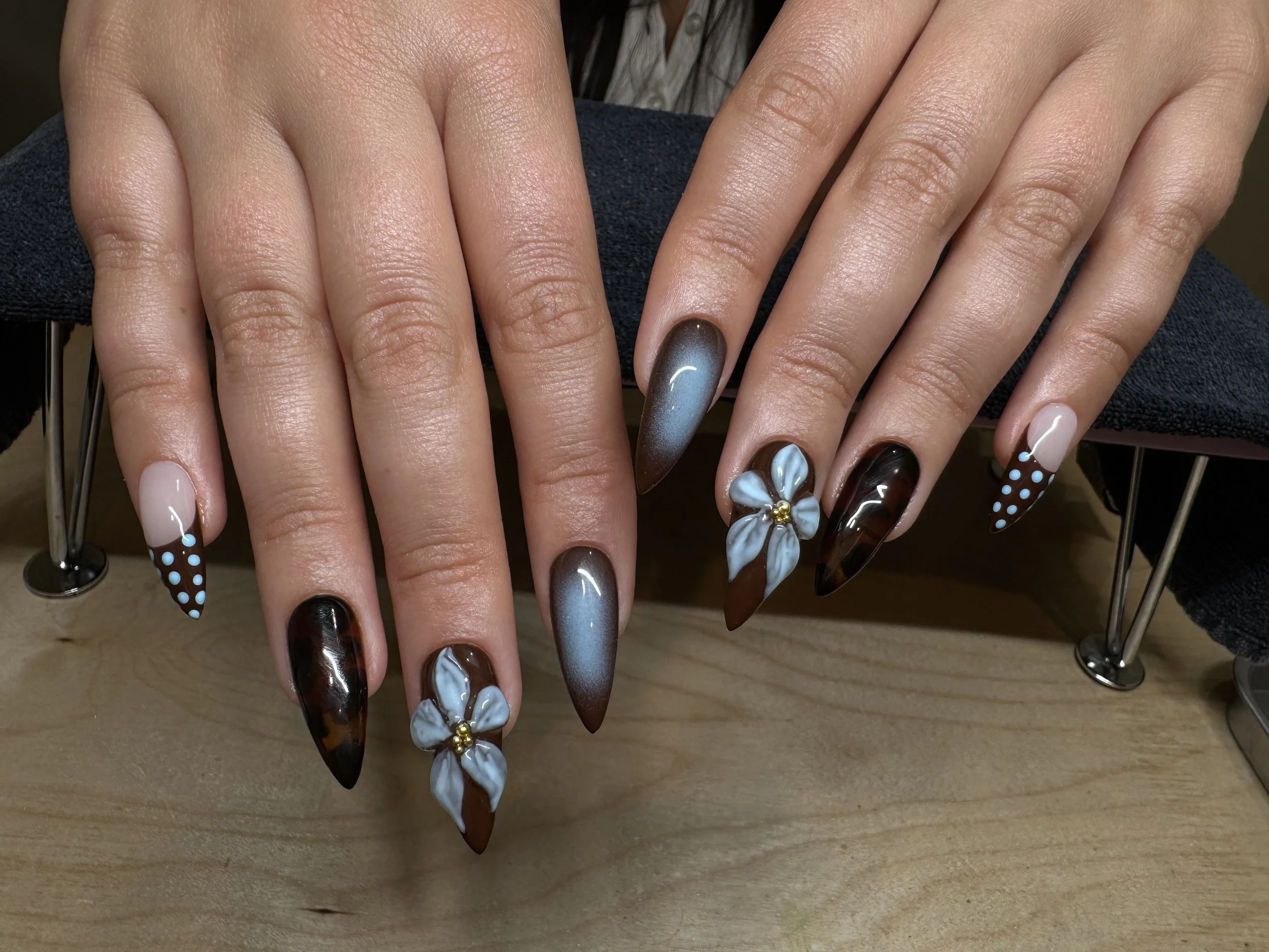 Hands with long, pointed nails painted in various shades of brown, blue, and white, some with floral and polka dot designs, resting on a wooden surface.