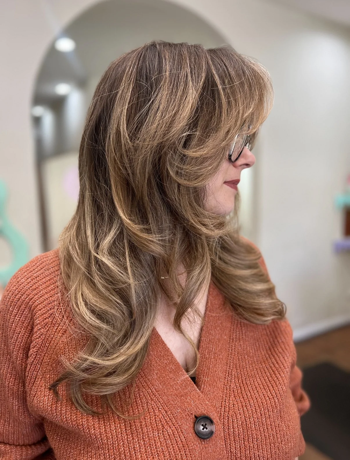 A woman with wavy, shoulder-length light brown hair and glasses, wearing an orange sweater, in profile view.