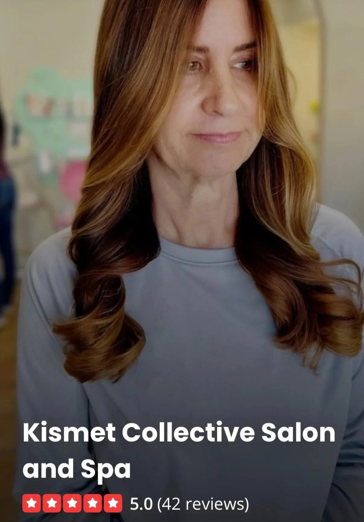 Close-up of a woman with long, wavy, reddish-brown hair wearing a light gray top, at Kismet Collective Salon and Spa, with a blurred background and a review rating of 5 stars based on 42 reviews.