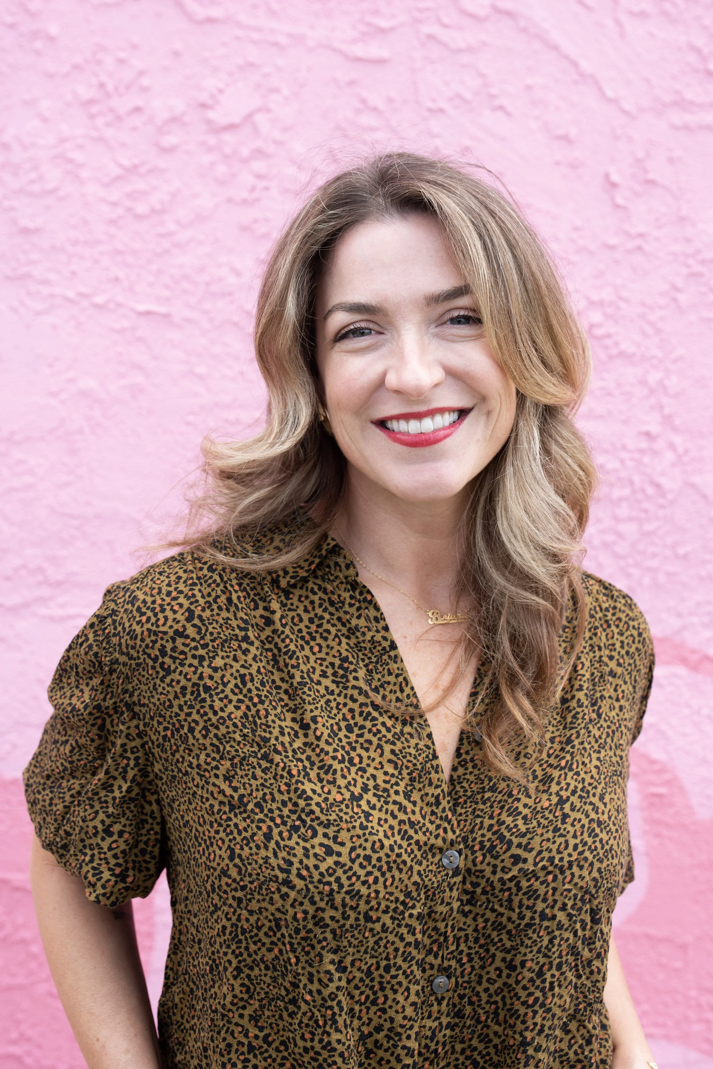 A woman with wavy, light brown hair, smiling, wearing a leopard print blouse, standing against a pink textured wall.
