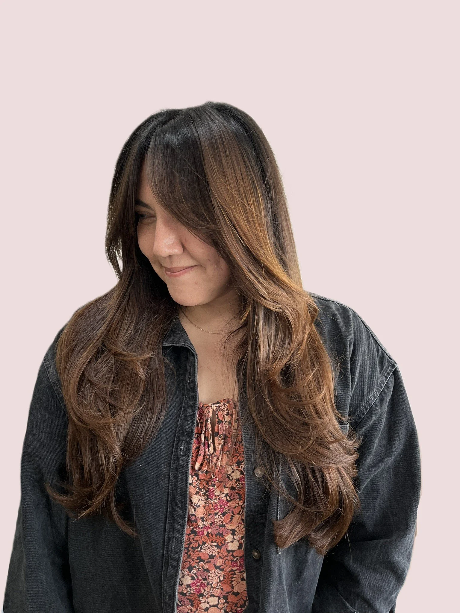 A woman with long, wavy hair that is dark at the roots and transitions to light brown and blonde at the ends, wearing a black denim jacket over a red and black floral dress, smiling softly and looking down slightly.