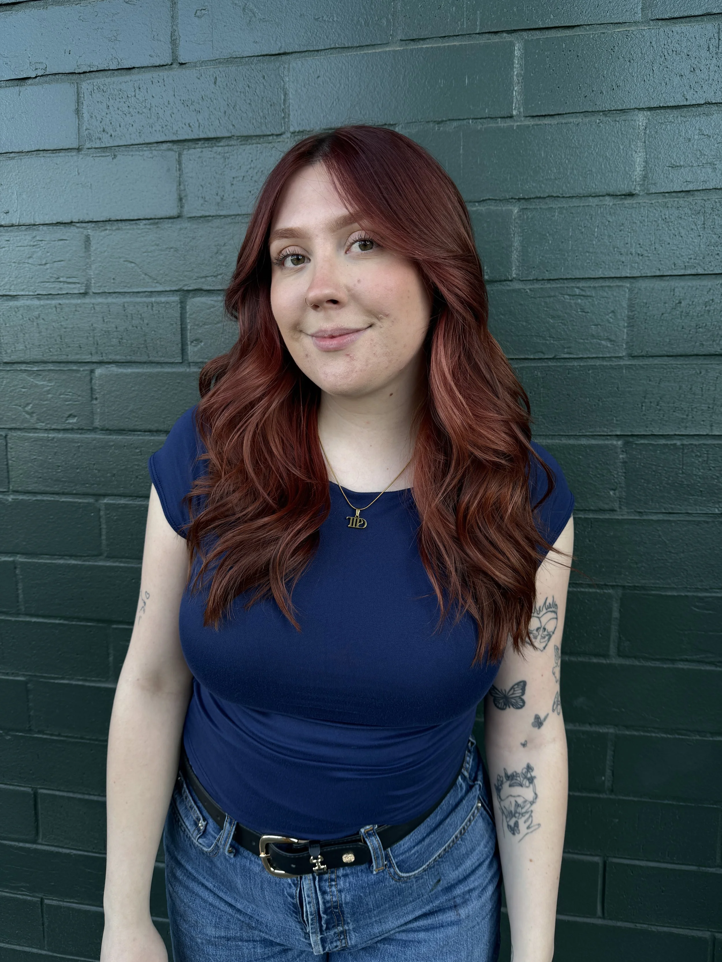 A young woman with long, wavy red hair wearing a navy blue top and blue jeans, standing in front of a green brick wall.