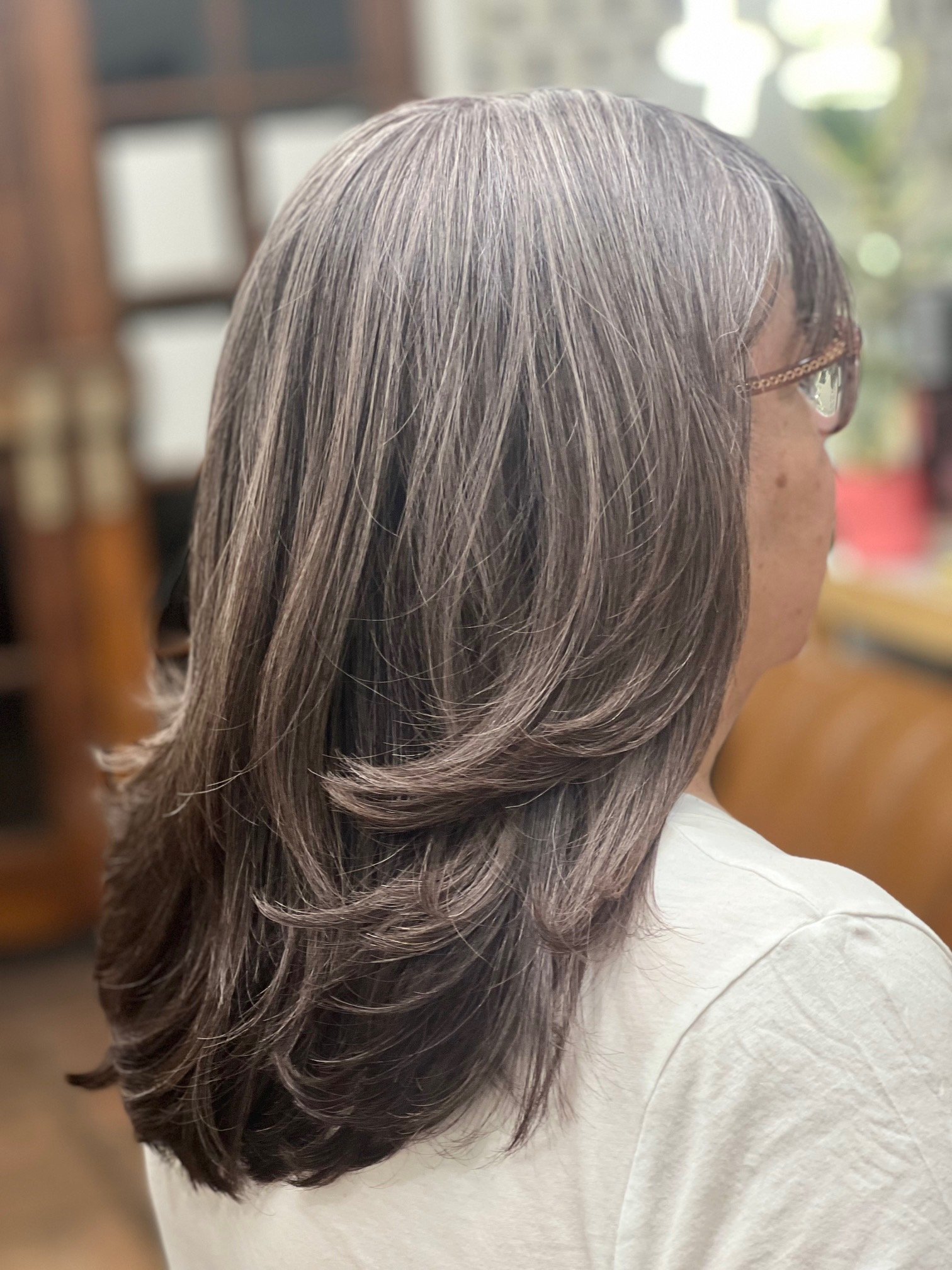 Side view of a woman with long, wavy salt-and-pepper hair, wearing glasses and a white shirt, sitting indoors.