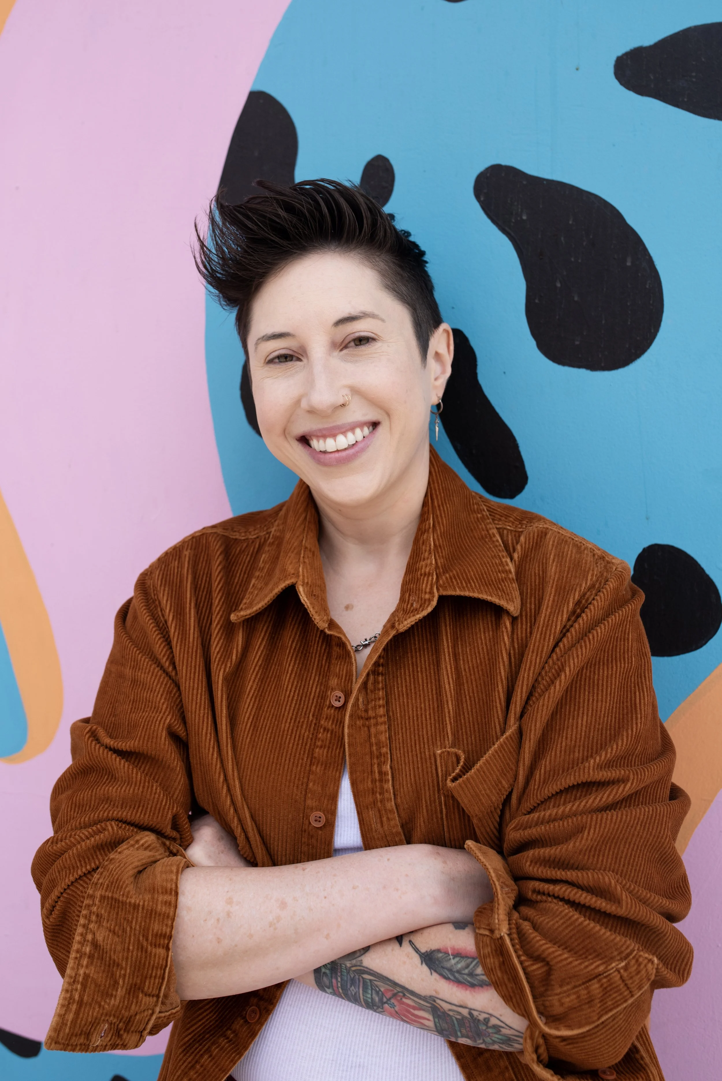 A person with short dark hair and tattoos on their arm, smiling with arms crossed, wearing a brown corduroy jacket against a colorful abstract background.