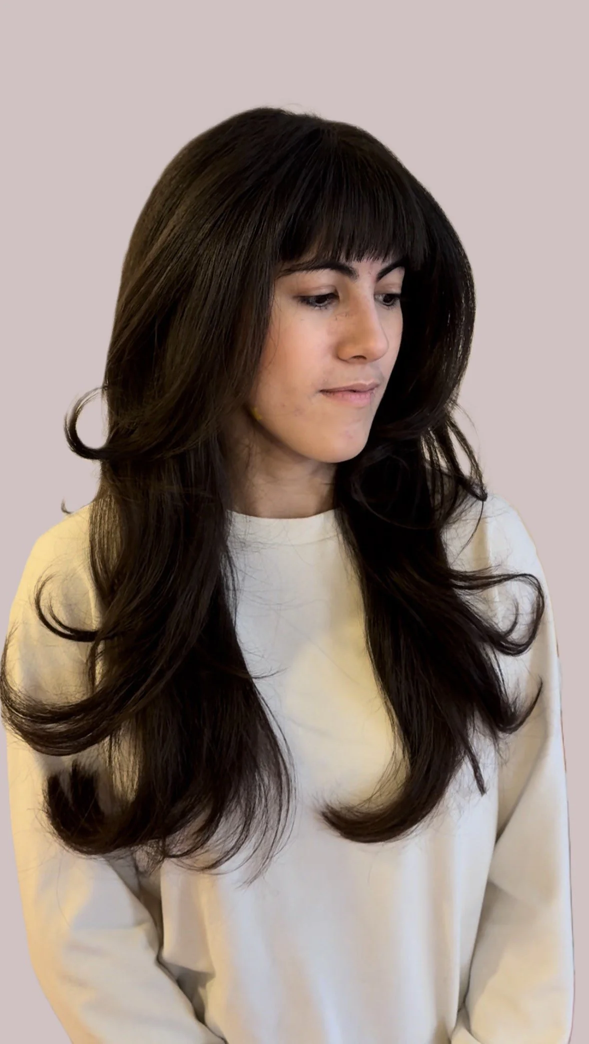 A woman with long, dark, wavy hair and bangs, wearing a light-colored top, looking downward against a plain background.