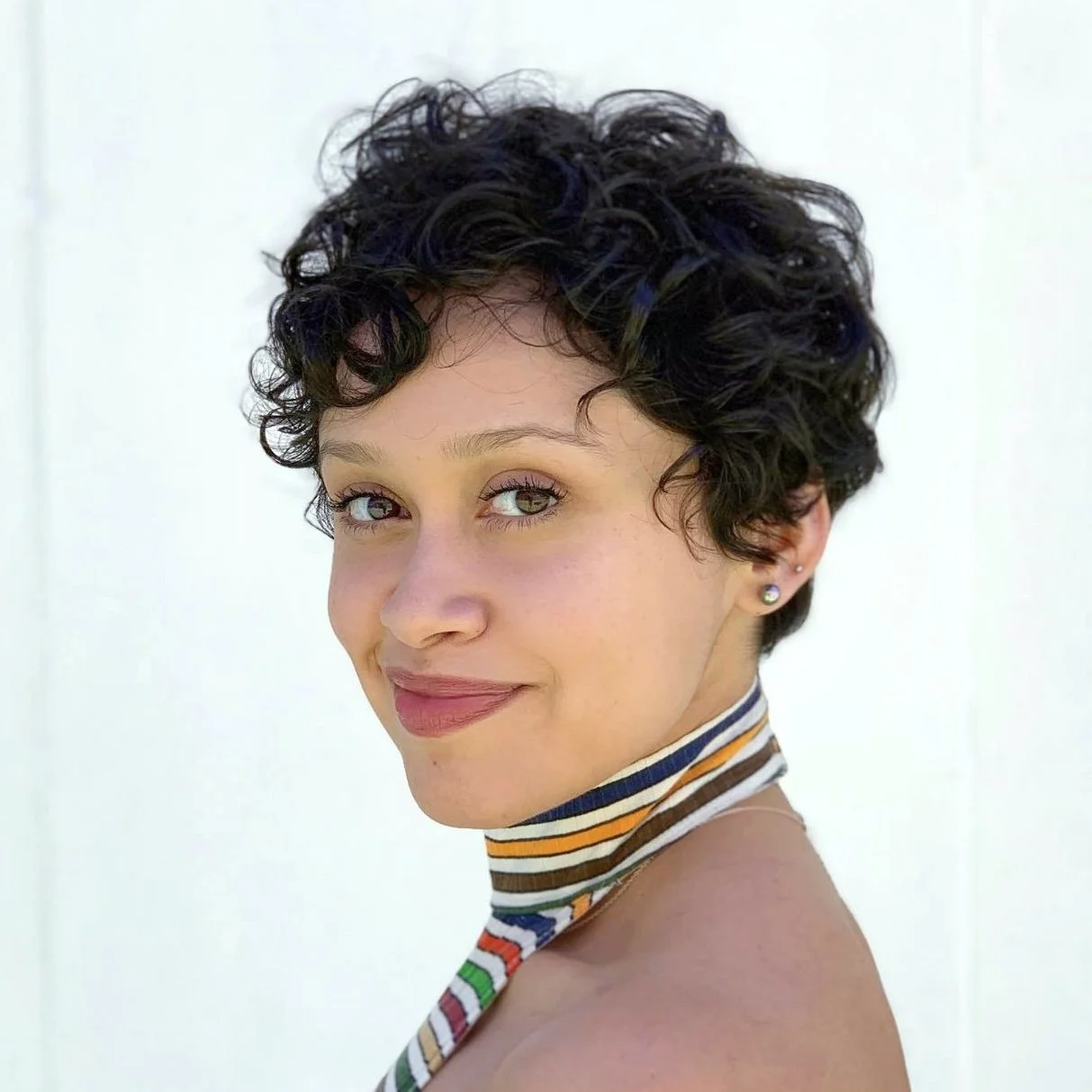 A woman with short, curly dark hair and green eyes, wearing earrings and a striped multicolored neck scarf, smiling against a white background.