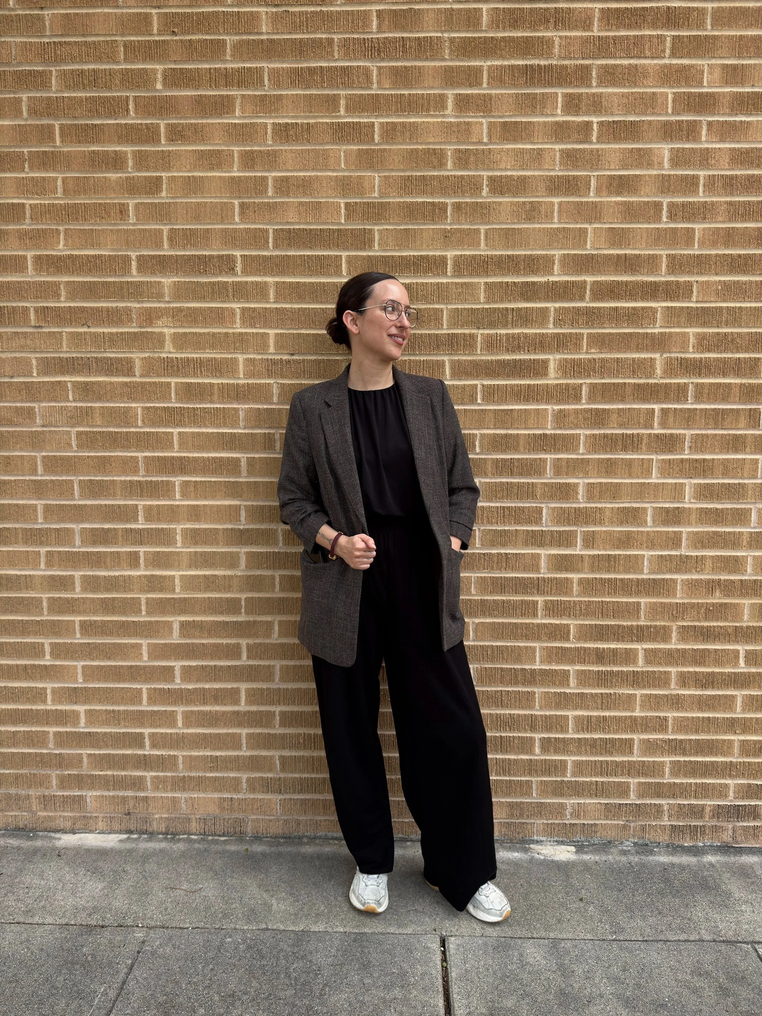 Julia Flaherty, a woman in glasses, a dark jacket, and black pants standing against a brick wall. She is smiling and looking to her left with her hands in her pockets. She is wearing white sneakers.