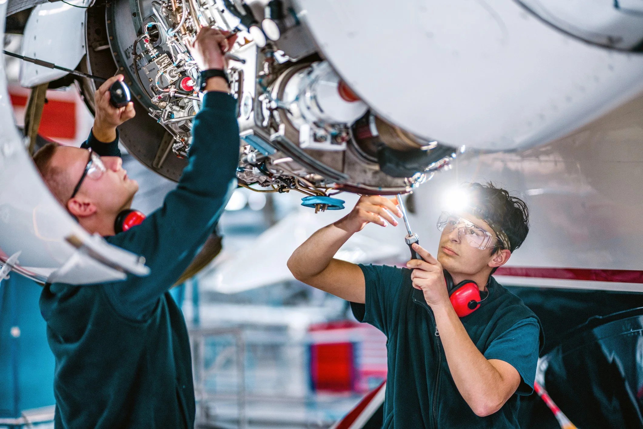 Two technicians working on an aircraft engine, one holding a screwdriver and the other adjusting components, in an aircraft maintenance hangar.