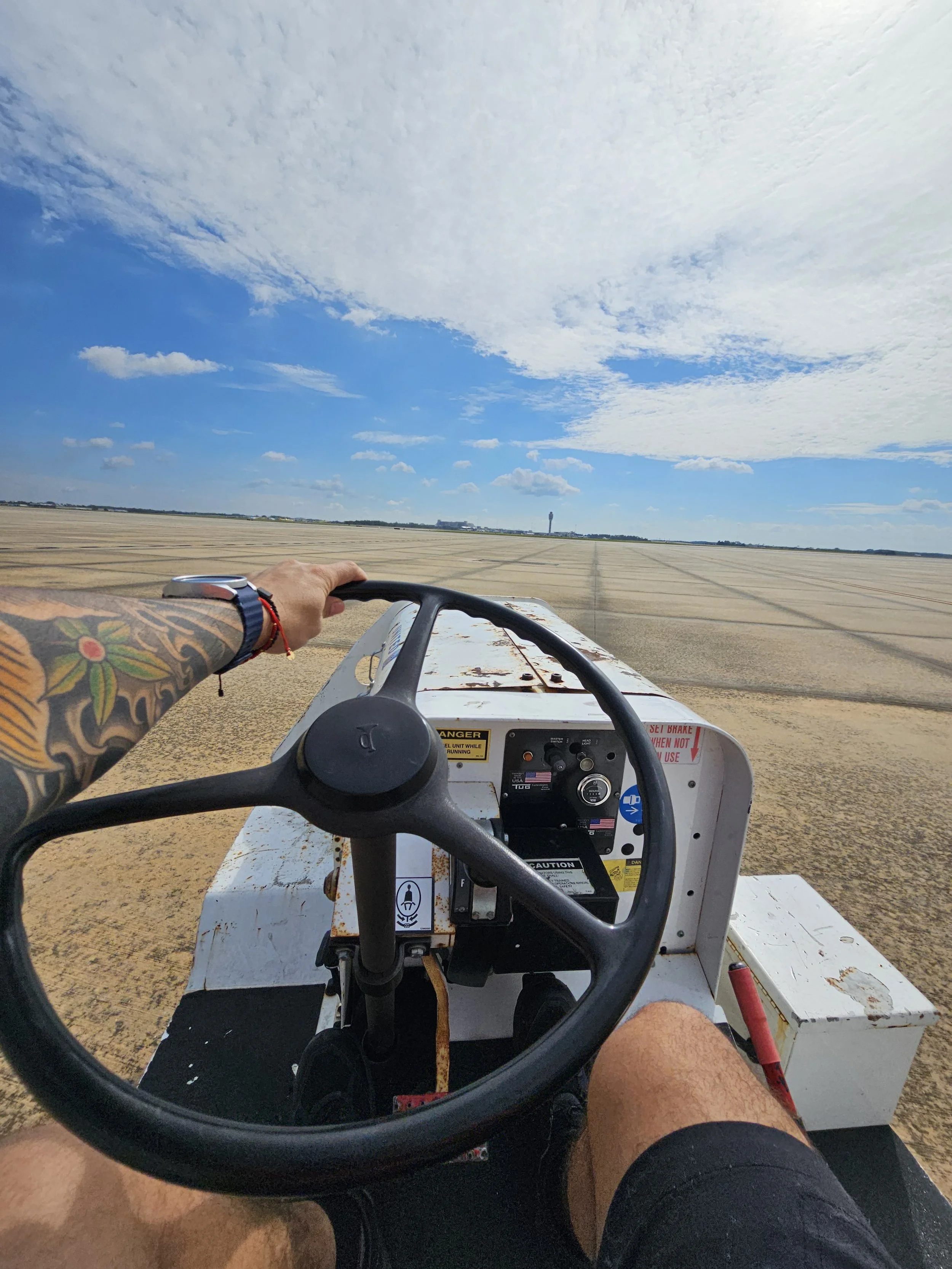 A person with a colorful tattoo on their arm driving a small white utility vehicle across a large open field of harvested farmland with a blue sky and scattered clouds overhead.