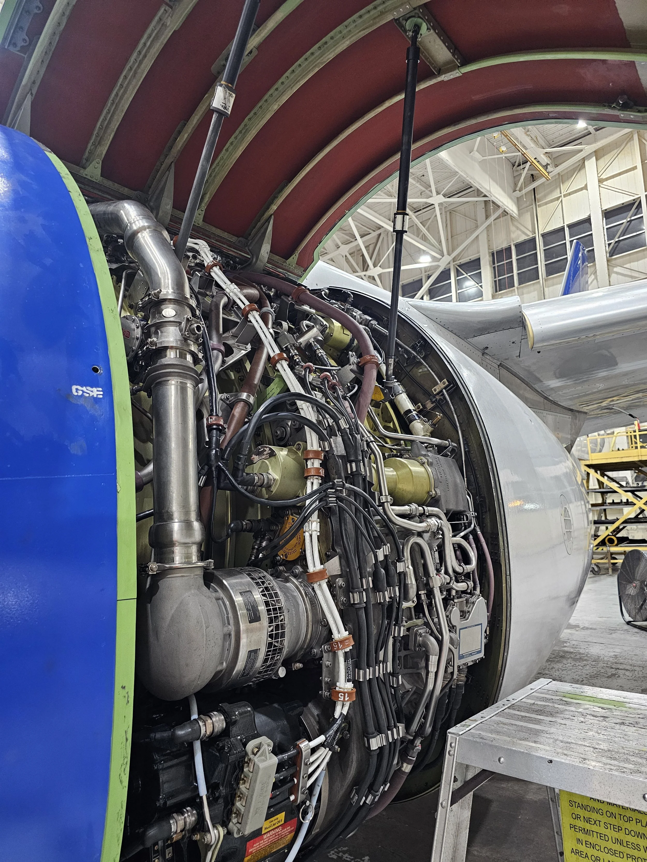 Close-up of an aircraft engine undergoing maintenance inside a hangar, showing various pipes, wires, and mechanical components.