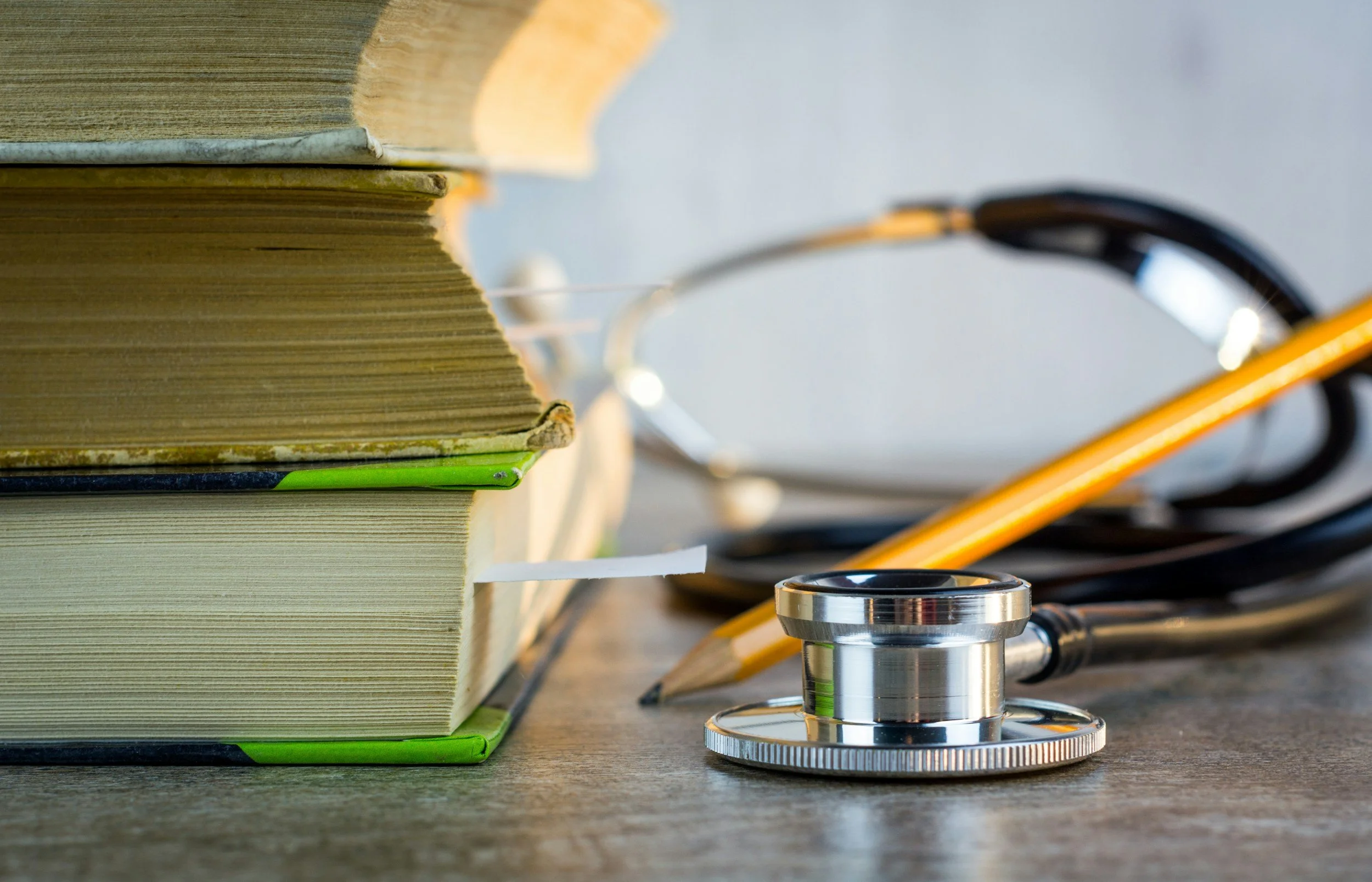 Books stacked on a table with a stethoscope and a yellow pencil nearby, indicating a medical or educational setting.